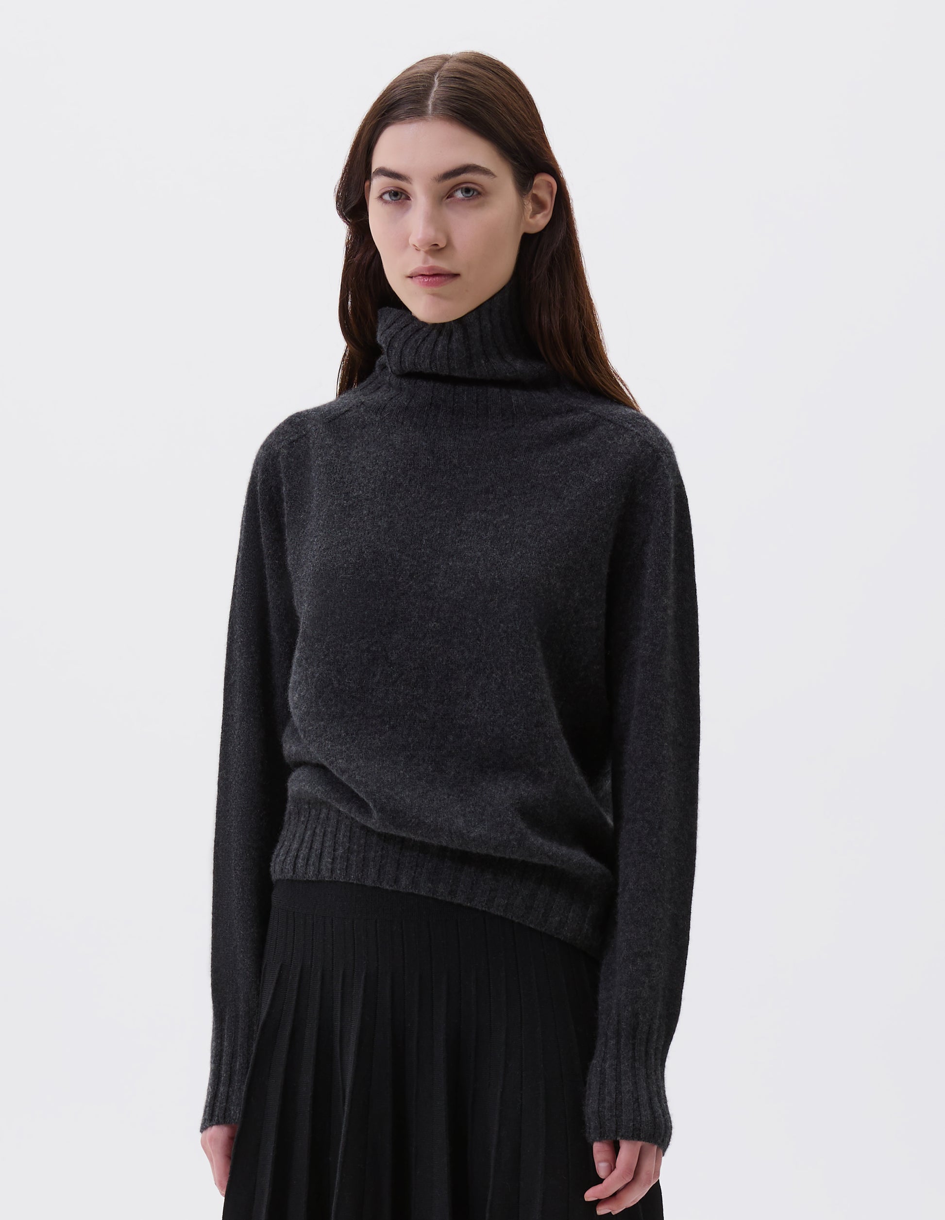 Cropped body image, front view of model wearing charcoal grey high neck cashmere jumper, with saddle sleeve and contrasting wide rib on neck, cuffs and hem. Styled with knitted black pleat skirt.