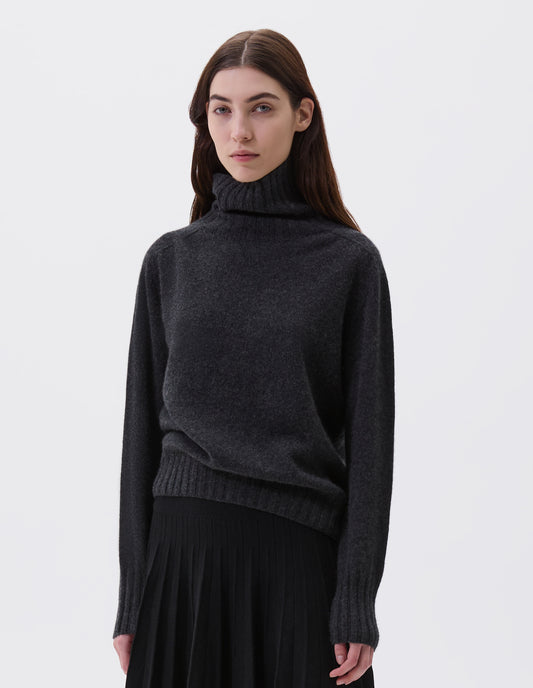 Cropped body image, front view of model wearing charcoal grey high neck cashmere jumper, with saddle sleeve and contrasting wide rib on neck, cuffs and hem. Styled with knitted black pleat skirt.
