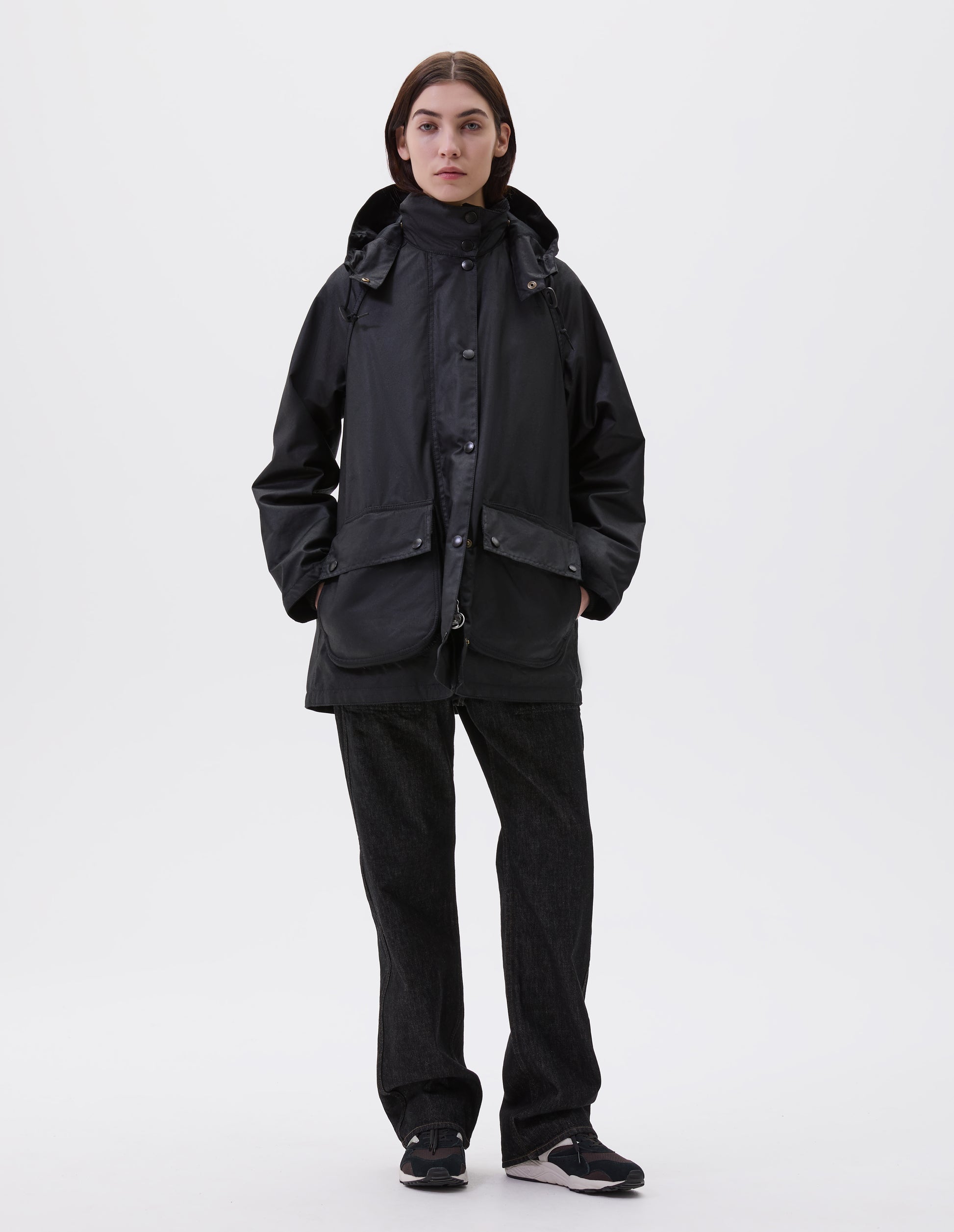 Full body image, front view of model wearing oversized black parka with stand collar and hood with wire adjustable peak, raglan sleeve, press stud fastening storm flap and large bellows pockets with press stud fastening envelope closure with concealed side pockets. Styled with black jeans and black, brown and white trainers.