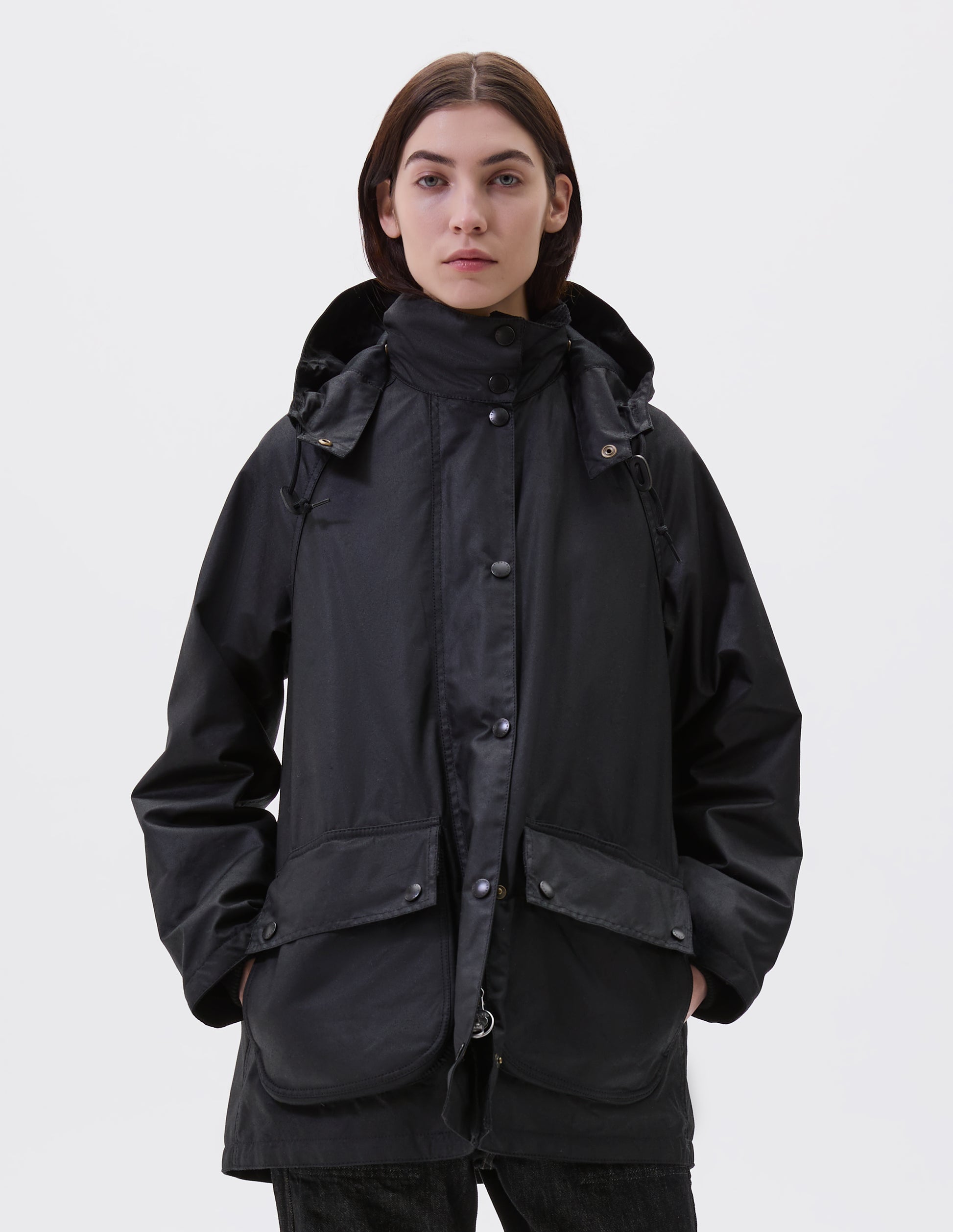 Cropped image, front view of model wearing oversized black parka with stand collar and hood with wire adjustable peak, raglan sleeve, large bellows pockets with press stud fastening envelope closure and concealed side pockets. Styled with black jeans.