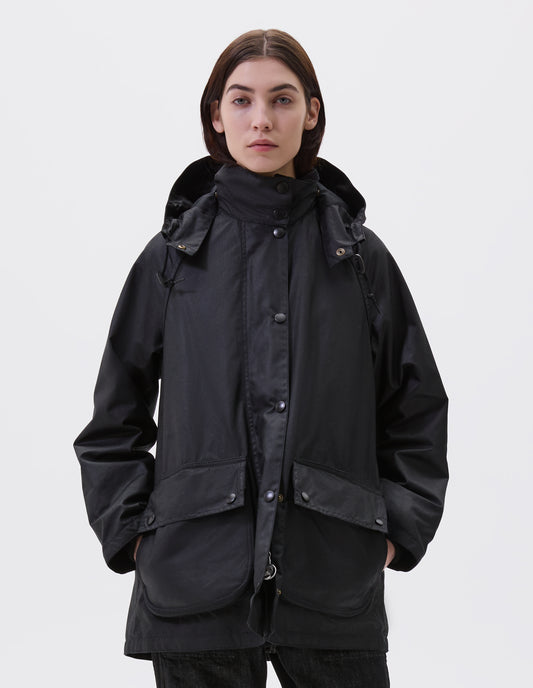 Cropped image, front view of model wearing oversized black parka with stand collar and hood with wire adjustable peak, raglan sleeve, large bellows pockets with press stud fastening envelope closure and concealed side pockets. Styled with black jeans.