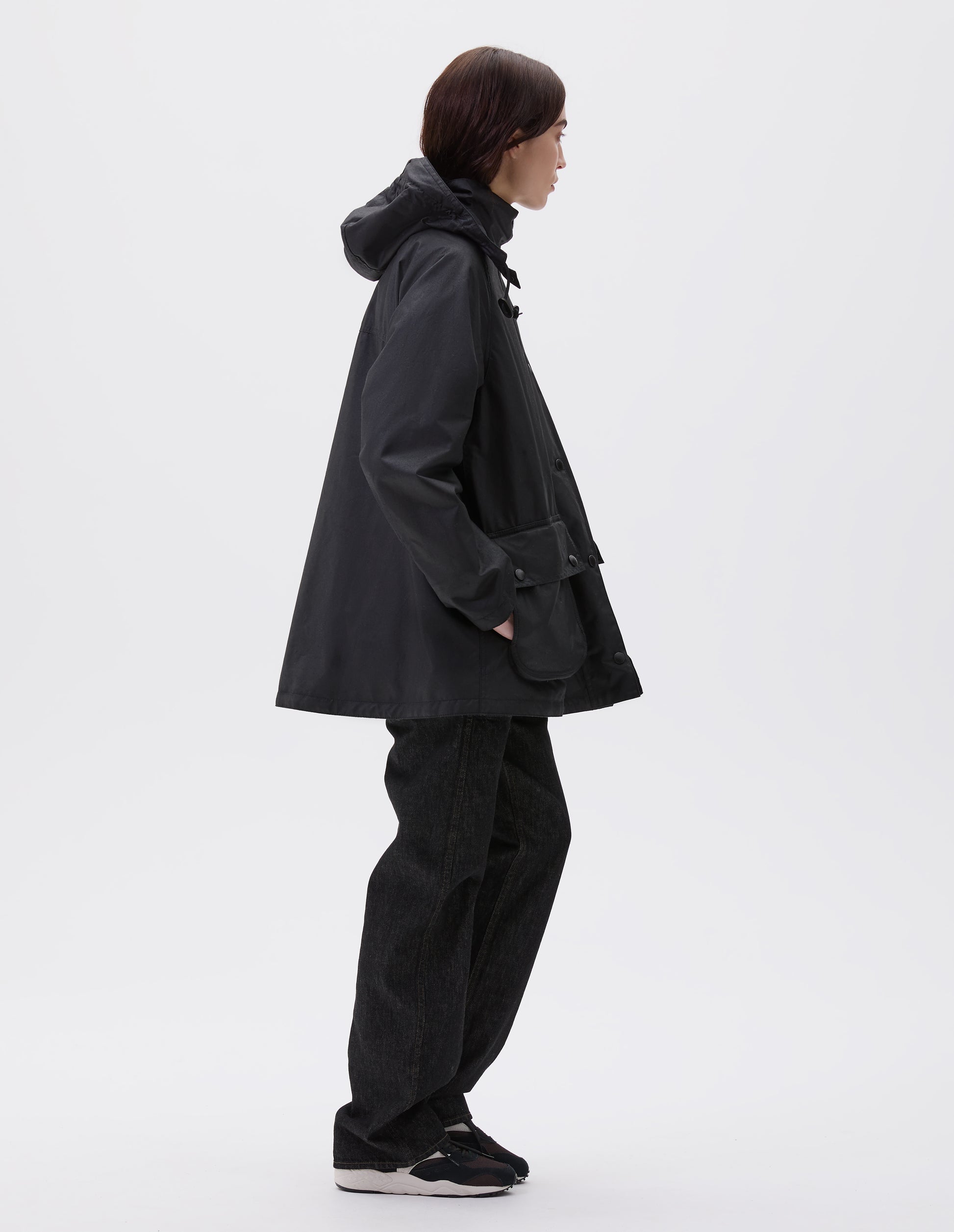 Full body image, side view of model wearing oversized black parka with stand collar and hood with wire adjustable peak, raglan sleeve, large bellows pockets with press stud fastening envelope closure and concealed side pockets. Styled with black jeans and black, brown and white trainers.