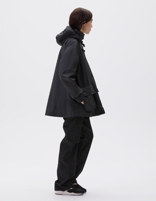 Full body image, side view of model wearing oversized black parka with stand collar and hood with wire adjustable peak, raglan sleeve, large bellows pockets with press stud fastening envelope closure and concealed side pockets. Styled with black jeans and black, brown and white trainers.