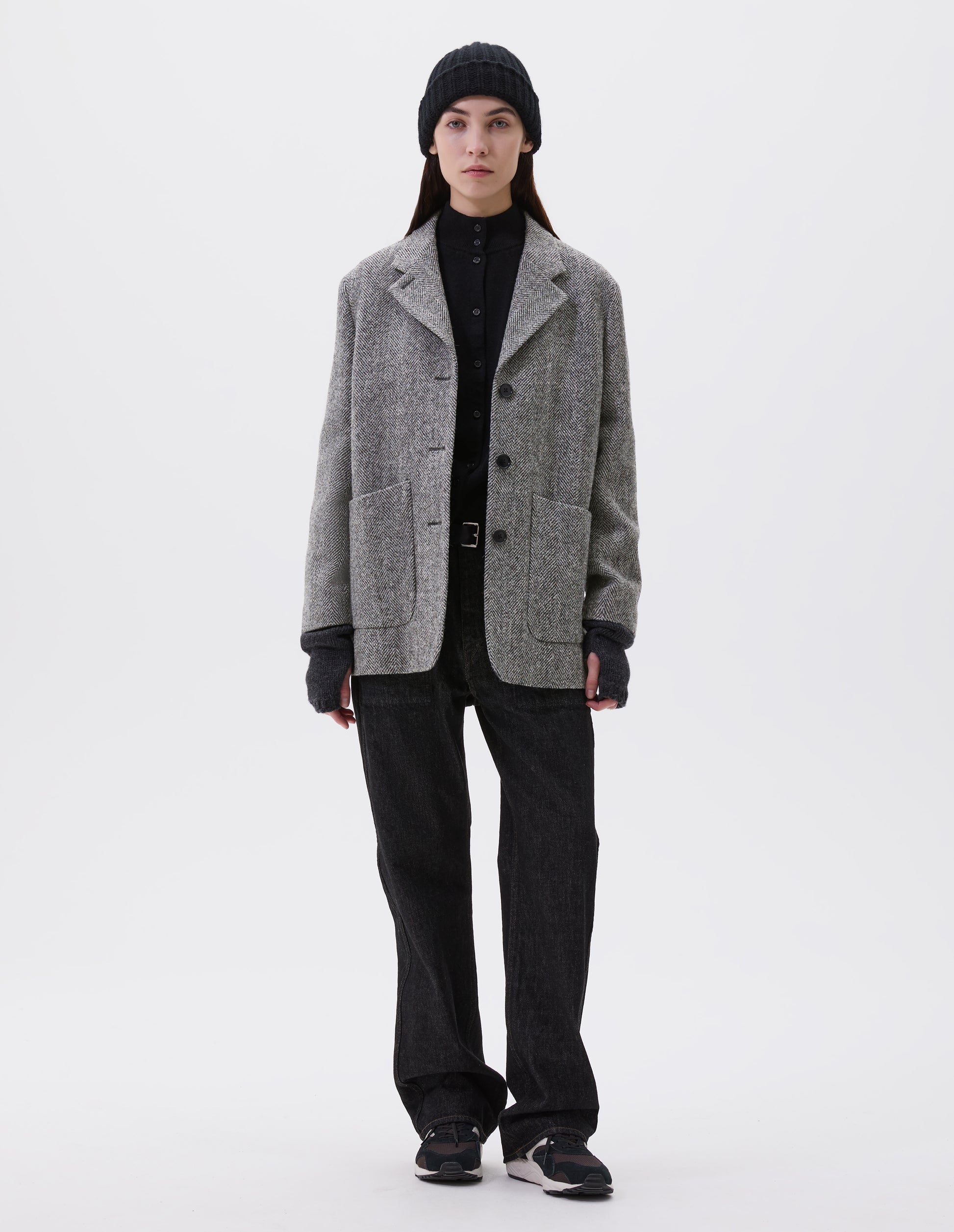 Full body image, front view of model wearing relaxed grey and black herringbone  blazer with horn buttons and large patch pockets. Styled with black beanie, black turtleneck, black jeans, black leather belt, dark grey wrist warmers and black, brown and white trainers.