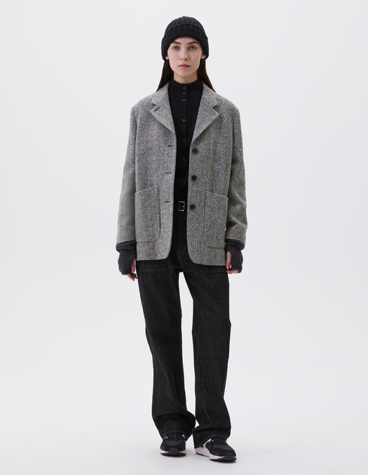 Full body image, front view of model wearing relaxed grey and black herringbone  blazer with horn buttons and large patch pockets. Styled with black beanie, black turtleneck, black jeans, black leather belt, dark grey wrist warmers and black, brown and white trainers.