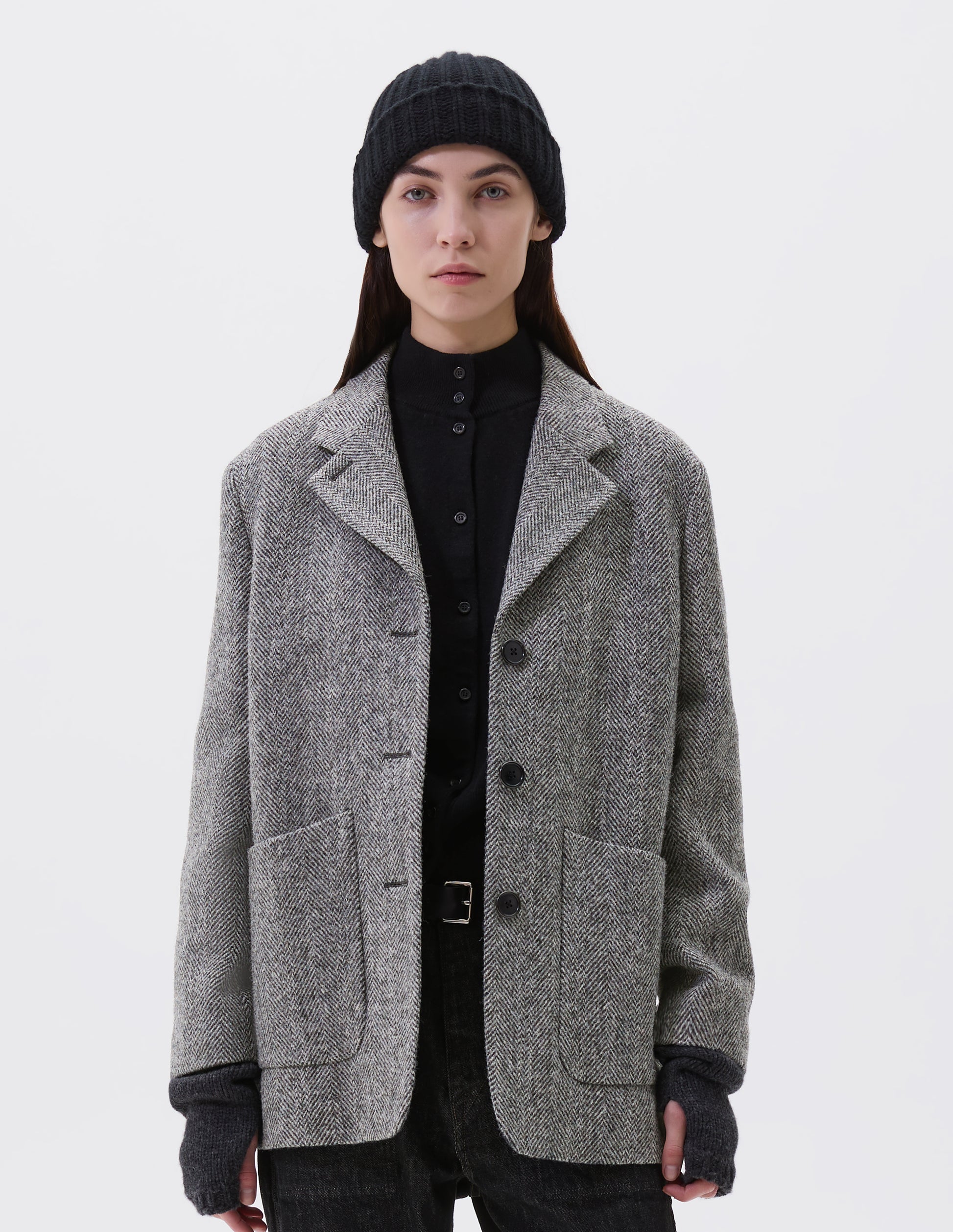 Cropped image, front view of model wearing relaxed grey and black herringbone  blazer with horn buttons and large patch pockets. Styled with black beanie, black turtleneck, black jeans, black leather belt and dark grey wrist warmers.