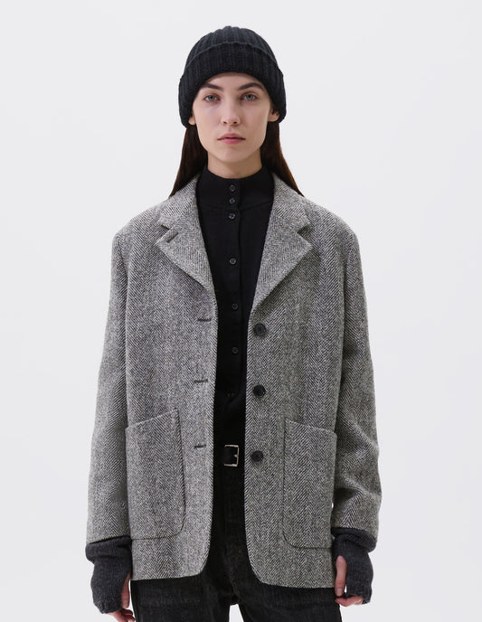 Cropped image, front view of model wearing relaxed grey and black herringbone  blazer with horn buttons and large patch pockets. Styled with black beanie, black turtleneck, black jeans, black leather belt and dark grey wrist warmers.