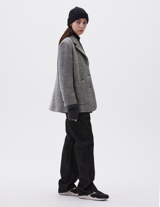 Full body image, side view of model wearing relaxed grey and black herringbone blazer with large patch pockets. Styled with black beanie, black turtleneck, black jeans, dark grey wrist warmers and black, brown and white trainers.