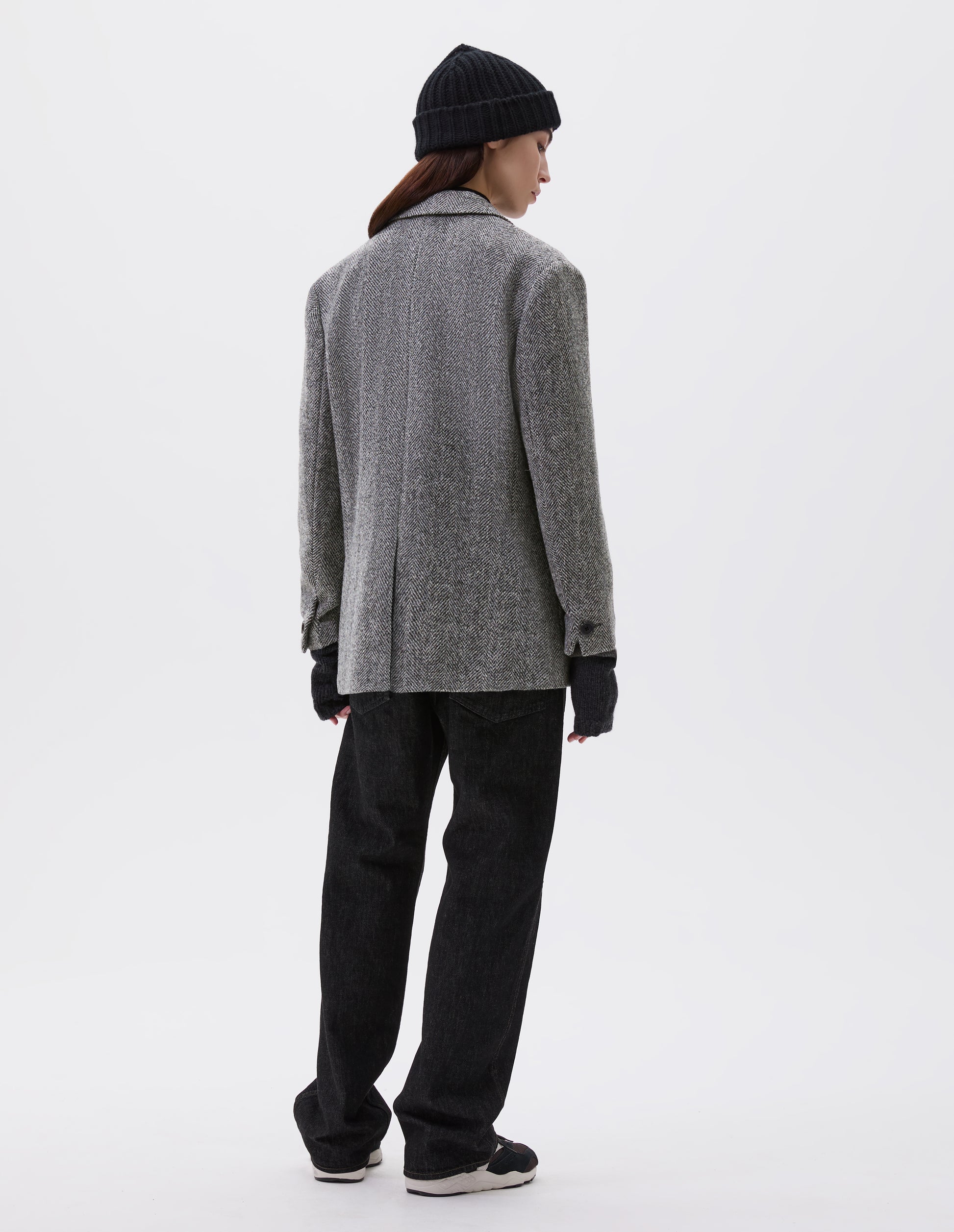 Full body image, back view of model wearing relaxed grey and black herringbone blazer with horn buttons on cuffs and back centre vent. Styled with black beanie, black jeans, dark grey wrist warmers and black, brown and white trainers.