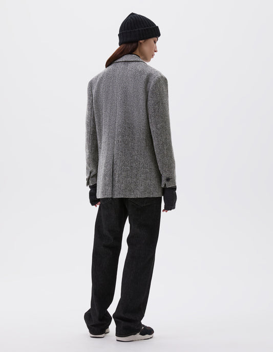 Full body image, back view of model wearing relaxed grey and black herringbone blazer with horn buttons on cuffs and back centre vent. Styled with black beanie, black jeans, dark grey wrist warmers and black, brown and white trainers.