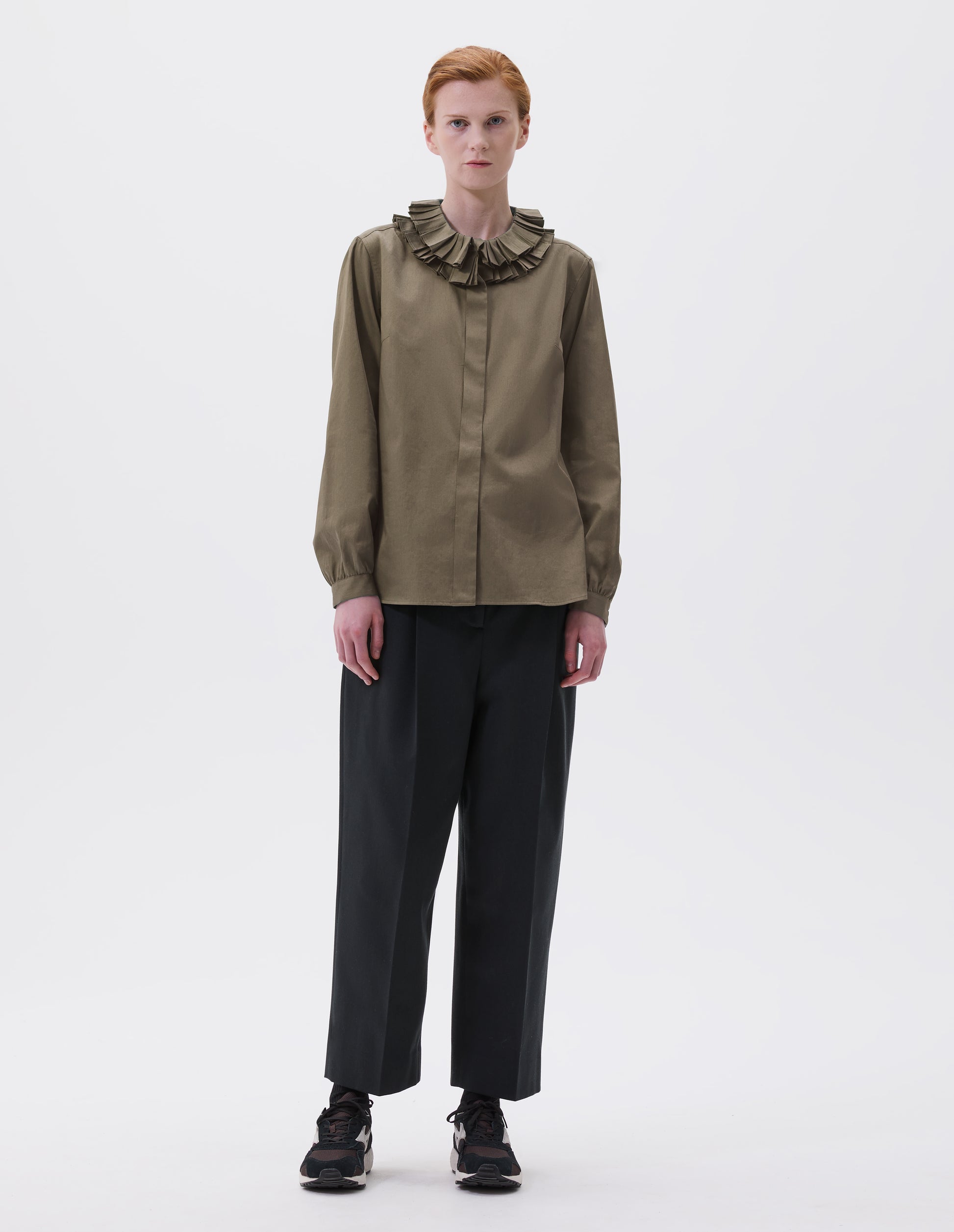 Full body image, front view of model wearing dark green long sleeve shirt with ruffled collar, concealed placket and shirt cuffs. Styled with cropped black trousers, black socks and black and brown trainers.