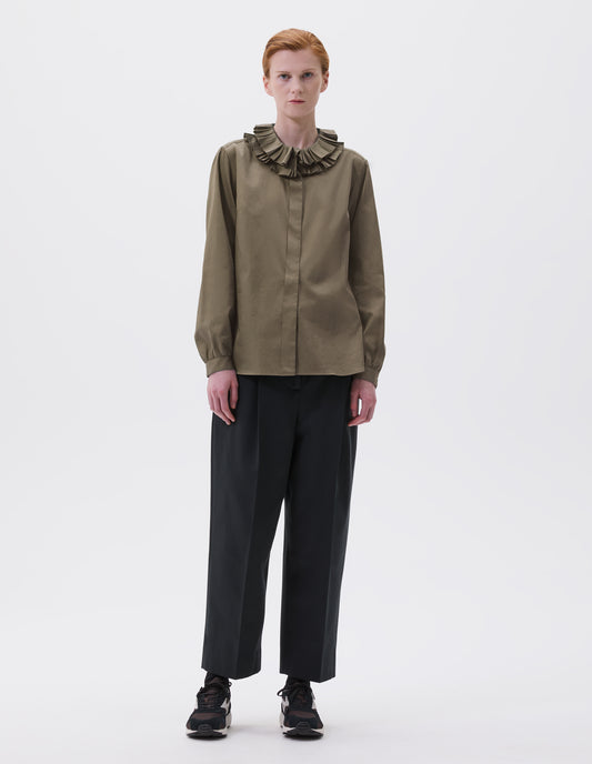 Full body image, front view of model wearing dark green long sleeve shirt with ruffled collar, concealed placket and shirt cuffs. Styled with cropped black trousers, black socks and black and brown trainers.