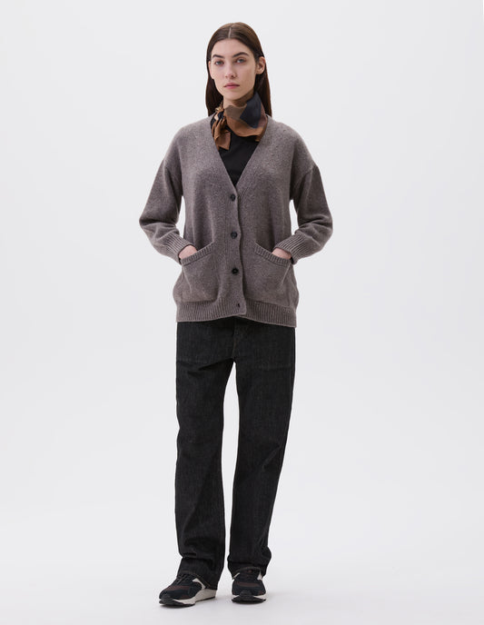 Full body image, front view of model wearing beige v-neck cardigan, with contrast rib on placket, cuffs and hem and two front pockets, finished with horn button closure. Styled with black t-shirt, multicoloured silk scarf, black denim jeans and black and brown trainers.