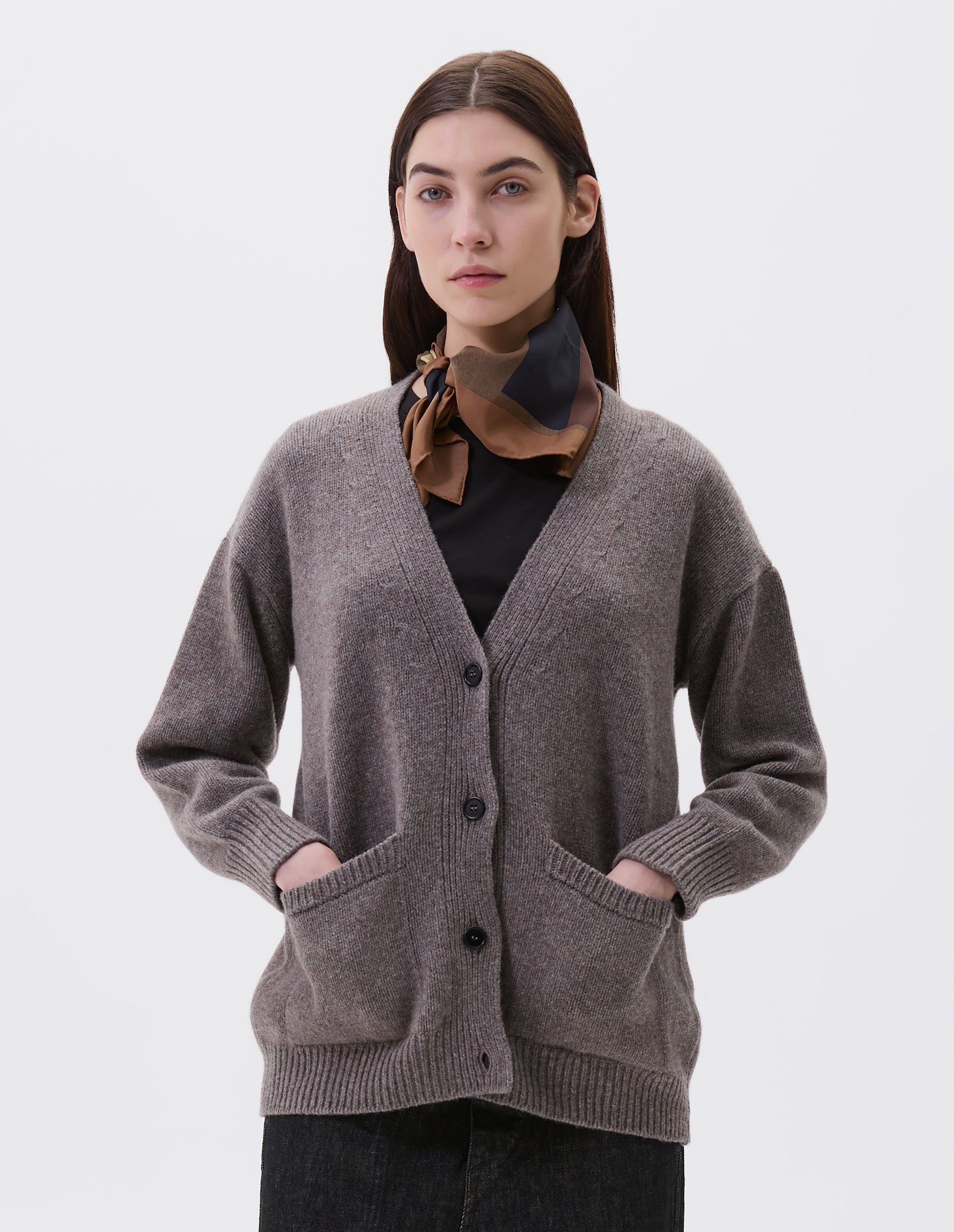 Cropped body image, front view of model wearing beige v-neck cardigan, with contrast rib on placket, cuffs and hem and two front pockets, finished with horn button closure. Styled with black t-shirt, multicoloured silk scarf and black denim jeans.