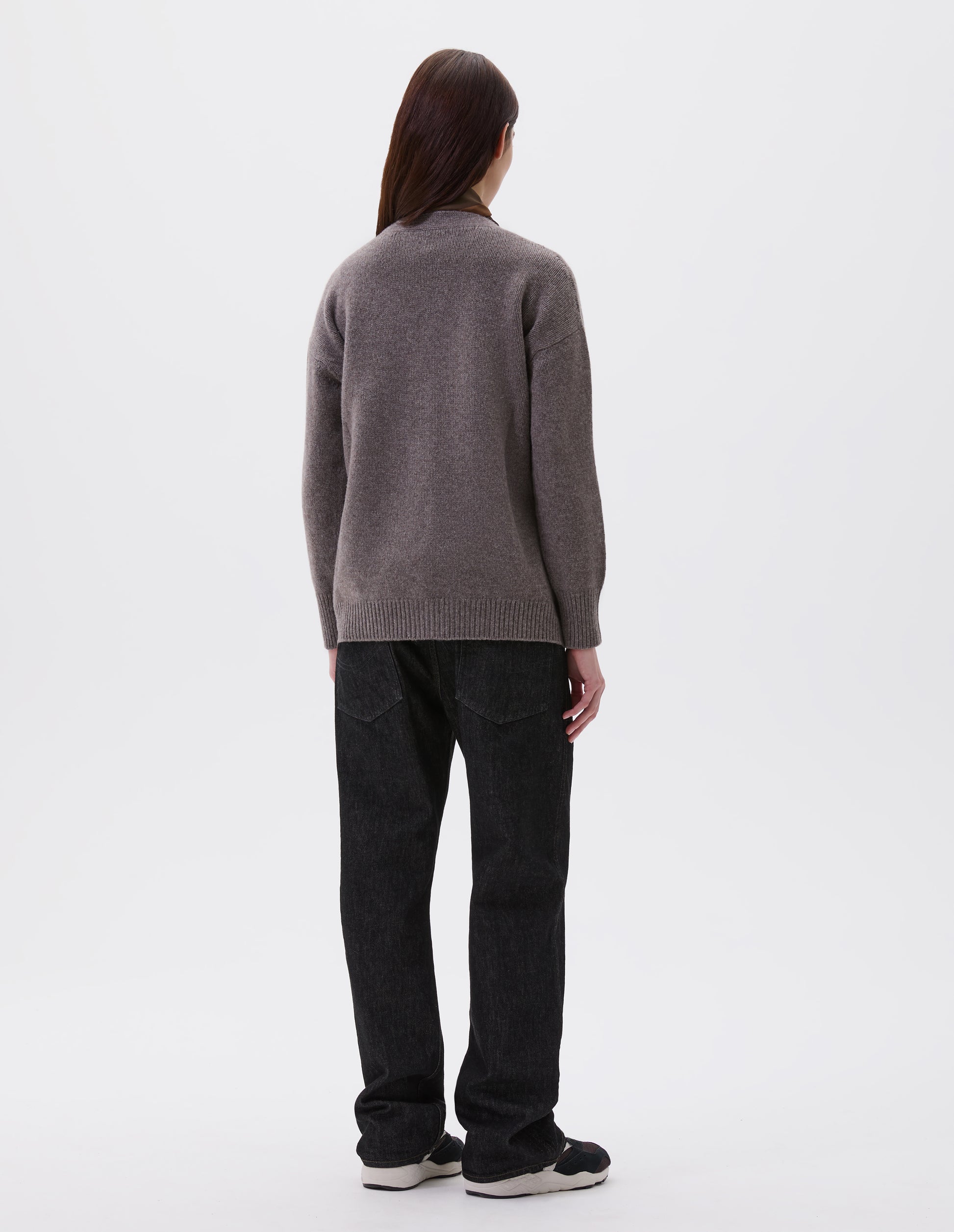 Full body image, back view of model wearing beige v-neck cardigan, with contrast rib cuffs and hem.  Styled with black denim jeans and black and brown trainers.