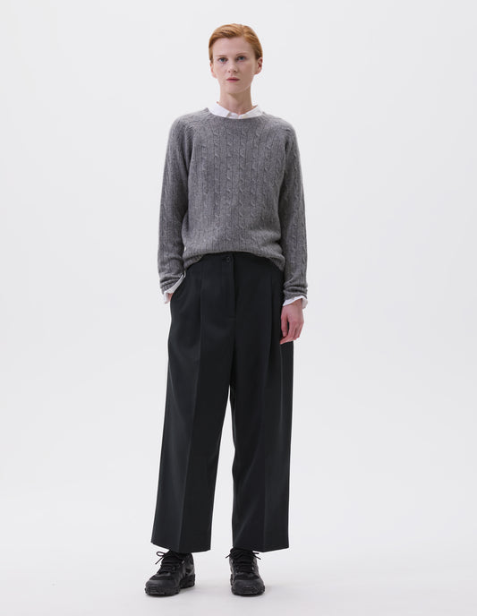 Full body image, front view of model wearing relaxed dark grey  cropped trousers with pleats and side pockets. Styled  light grey jumper over white shirt and black trainers.  