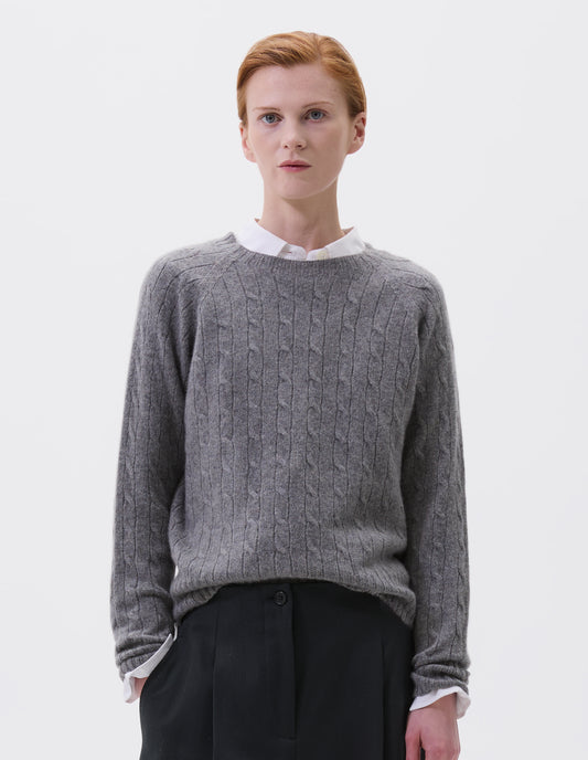 Cropped body image, front view of model wearing grey cable knit long sleeve jumper with raglan sleeve and contrast rib on neck, cuffs and hem. Styled with black cropped trousers and white shirt.