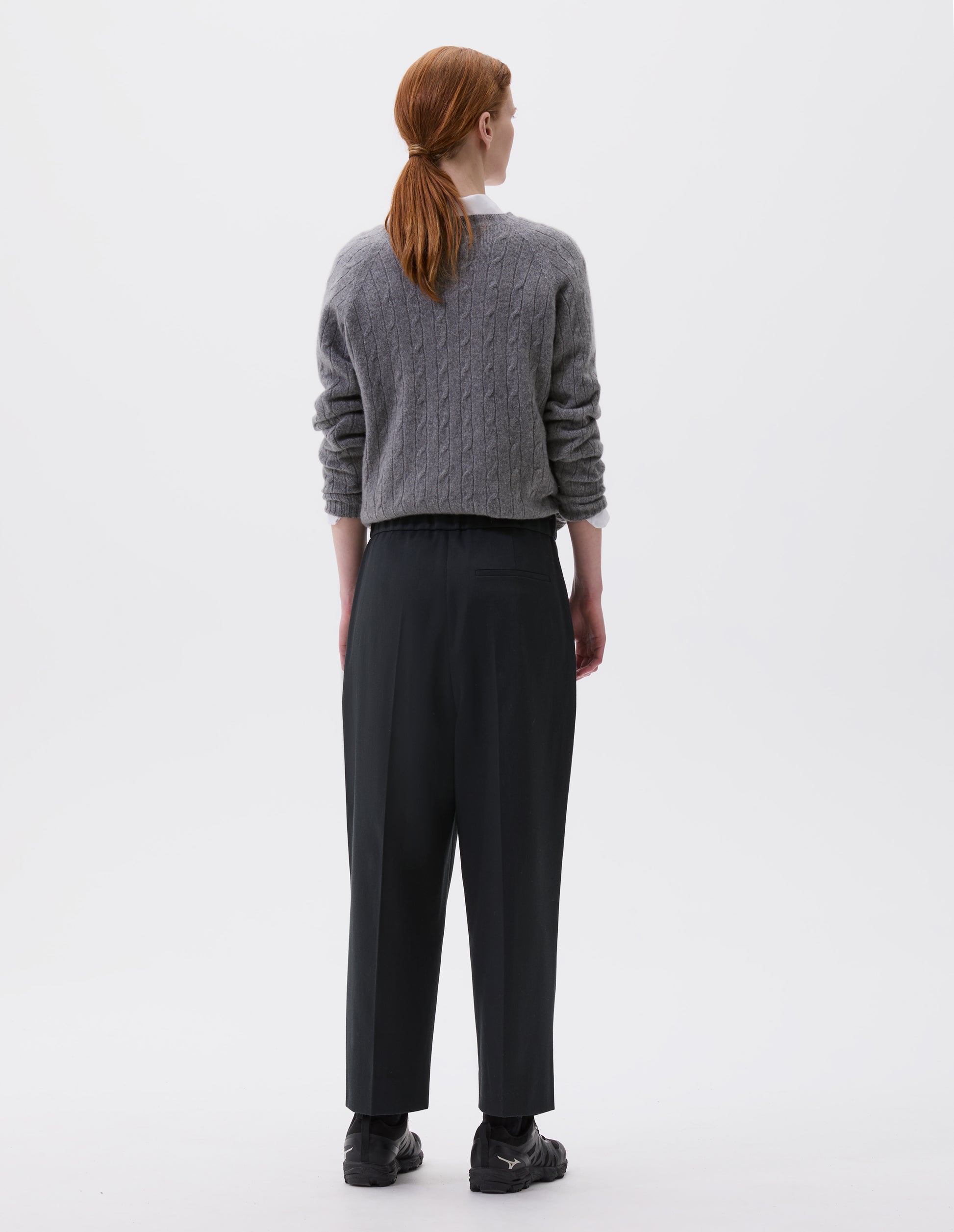 Full body image, back view of model wearing relaxed dark grey cropped trousers with pleats, half-elasticated waistband and welt back pocket. Styled with light grey jumper over white shirt and black trainers. 