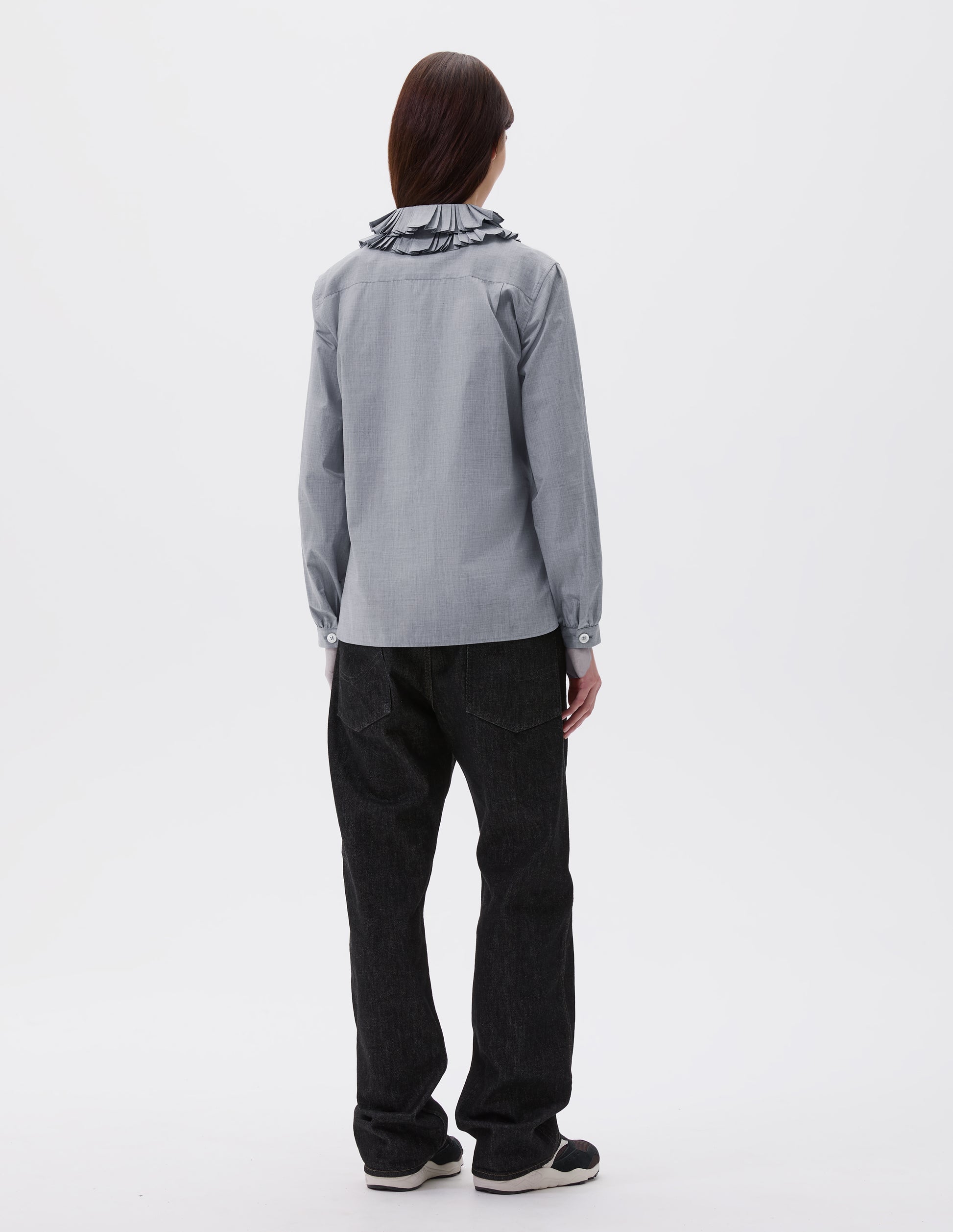 Full body image, back view of model wearing grey long sleeve shirt with ruffled collar and shirt cuffs. Styled with black denim jeans and black and brown trainers.