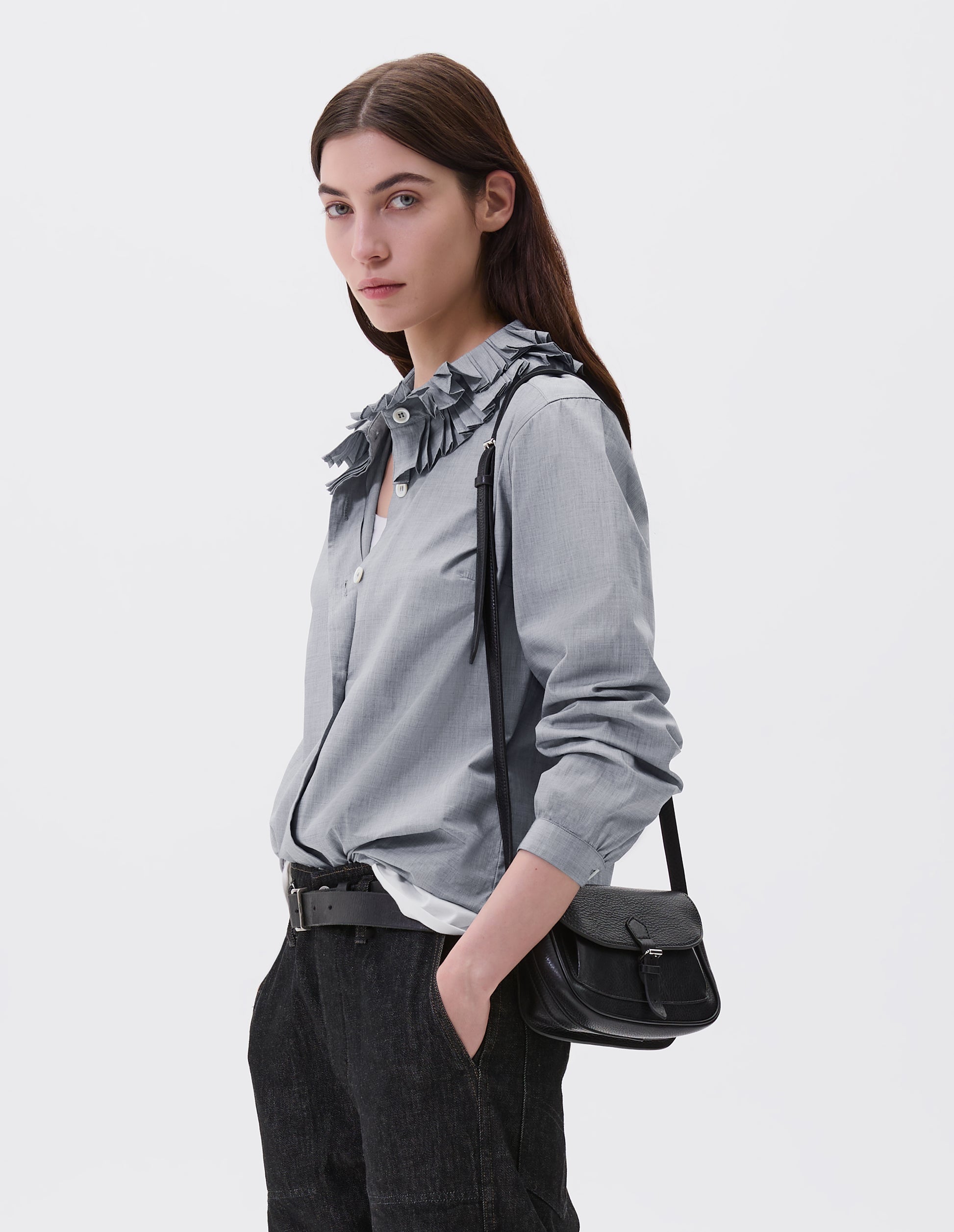 Cropped body image, front view of model wearing black fine grain leather small satchel bag with pleat expandable exterior pocket, roller buckle fastening closure and adjustable shoulder strap. Styled with grey collared shirt, black leather belt and black denim jeans.