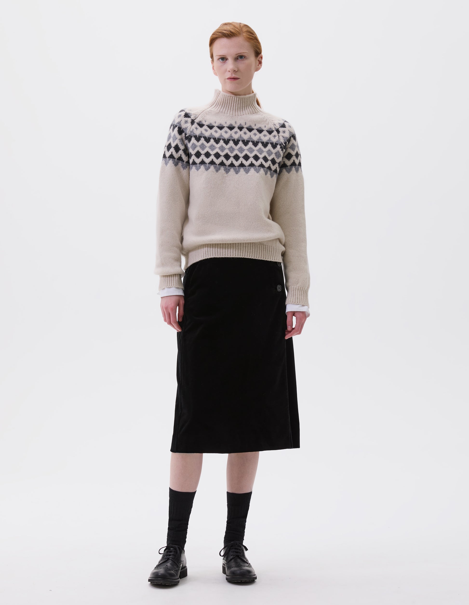 Full body image, front view of model wearing ecru and grey fair isle jumper with inset sleeve and contrasting rib on high neck, cuffs and hem. Styled over white long sleeve, with black velvet skirt, black socks and black leather brogues. 