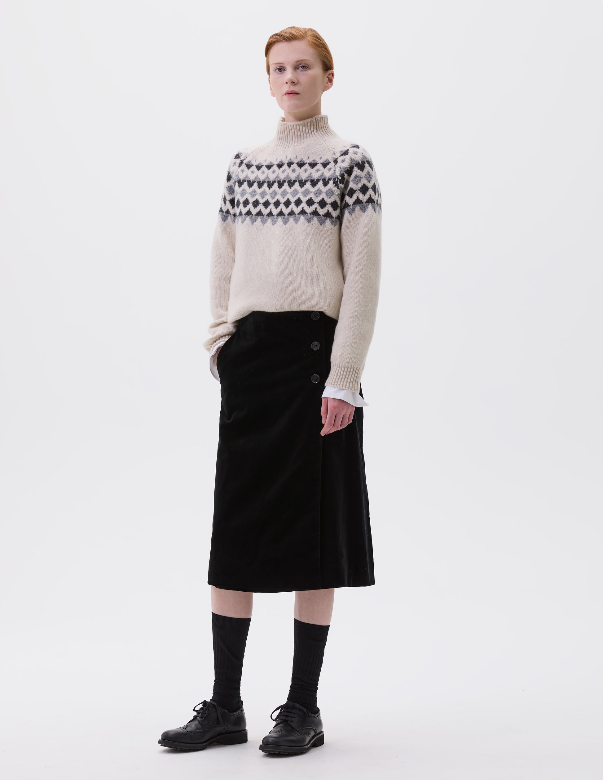 Full body image, front view of model wearing black velvet a-line skirt with deep wrap, horn button fastening and slant side pockets. Styled with cream and grey fair isle print jumper, black socks and black leather brogues.