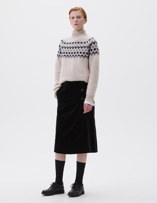 Full body image, front view of model wearing black velvet a-line skirt with deep wrap, horn button fastening and slant side pockets. Styled with cream and grey fair isle print jumper, black socks and black leather brogues.