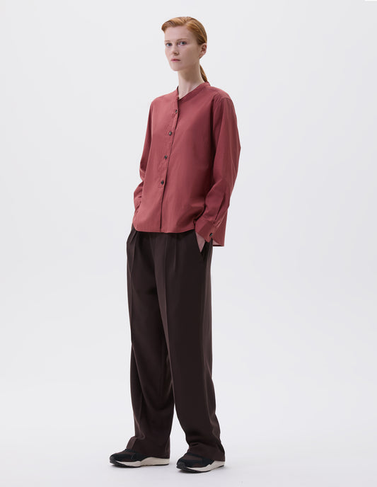 Full body image, front view of model wearing red pink long sleeve collarless shirt with raised placket and horn button closure and cuffs. Styled with brown pleated trousers and black and brown trainers.  