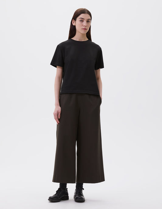 Full body image, front view of model wearing black cotton short sleeve t-shirt with boxy cut. Styled with black culottes, black socks and black leather brogues.