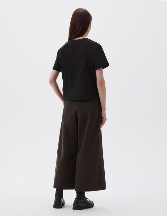 Full body image, back view of model wearing black cotton short sleeve t-shirt with boxy cut. Styled with black culottes, black socks and black leather brogues.