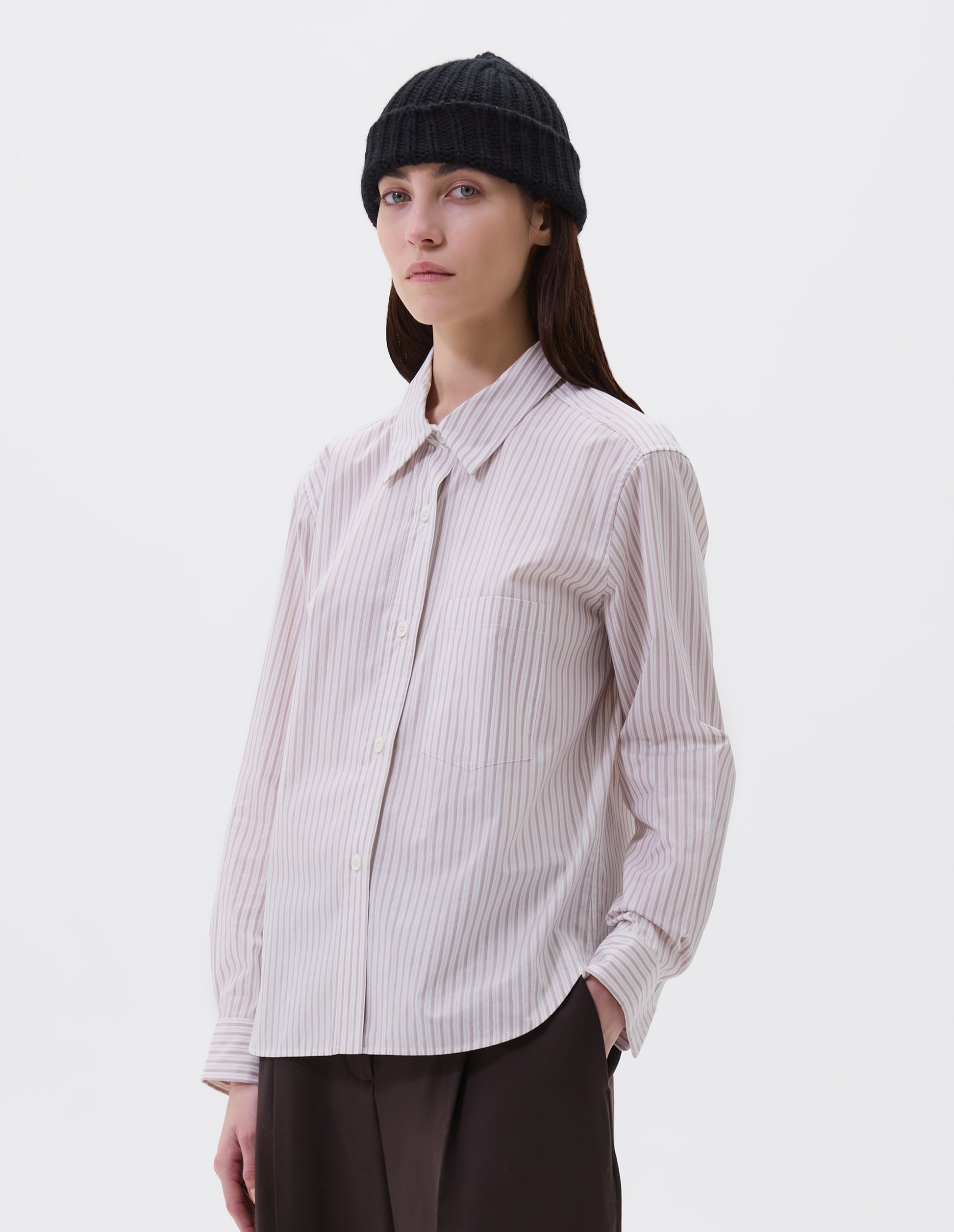 Cropped image, front view of model wearing beige, pink and black striped shirt with raised placket and mother of pearl buttons, patch pocket on chest. Styled with black beanie and dark brown trousers.