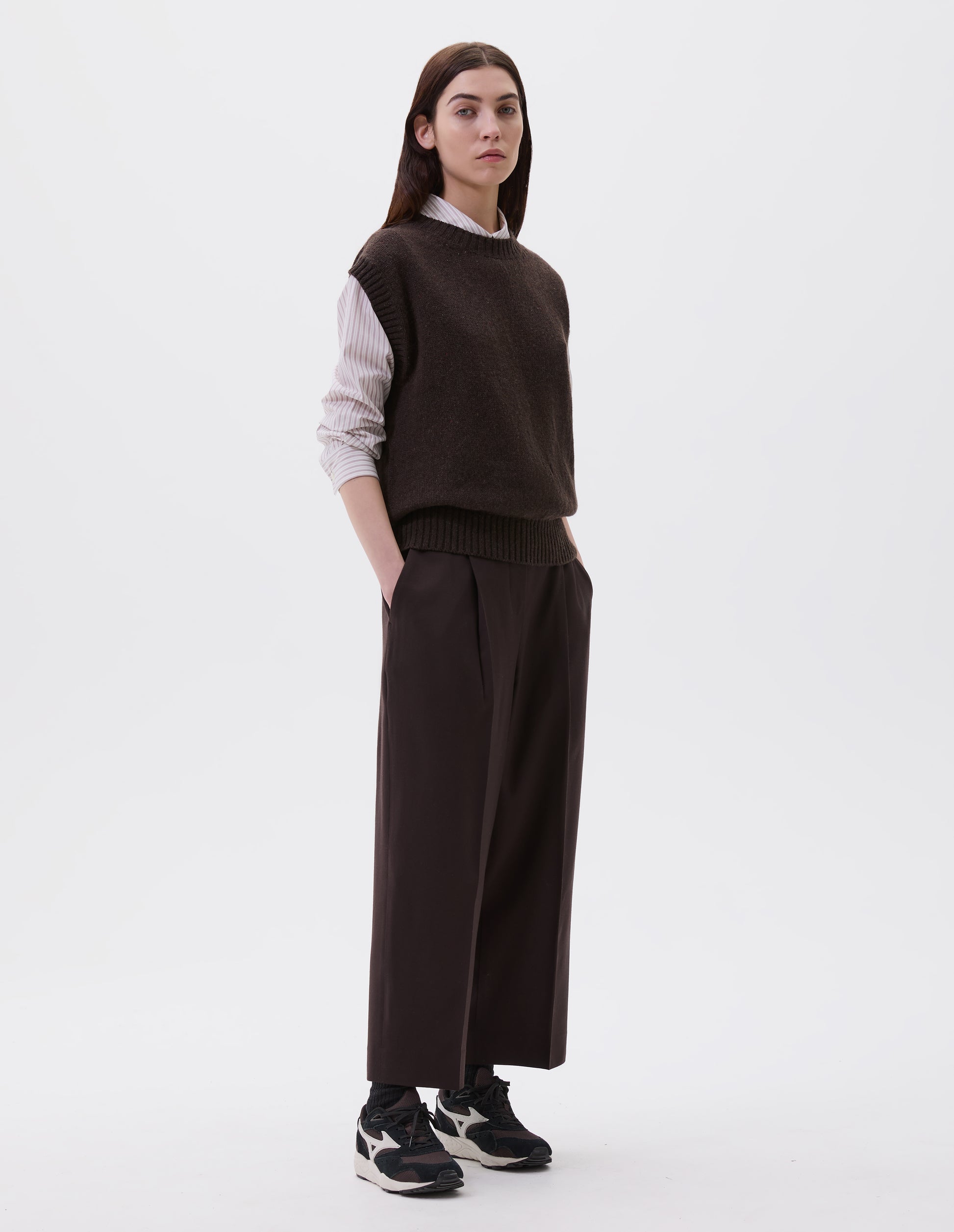 Full body image, front view of model wearing dark brown relaxed boxy slipover with ribbed neck, armhole and hem. Styled with white and pink striped shirt, dark brown trousers and black, brown and white trainers.