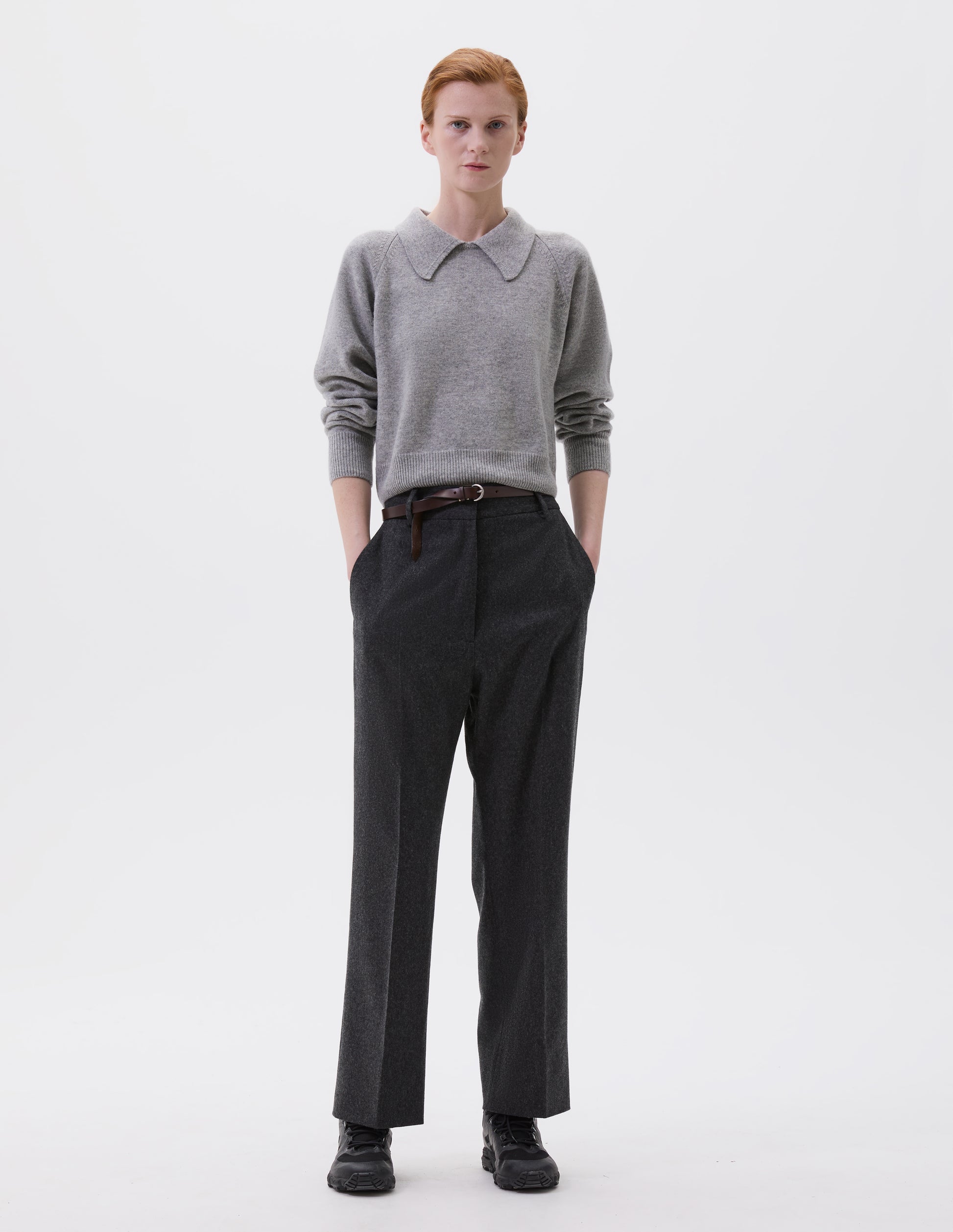 Full body image, front view of model wearing charcoal grey wool trousers with slant side pockets, belt loops and concealed zip fastening. Styled with grey collared jumper, brown slim leather belt and black trainers.