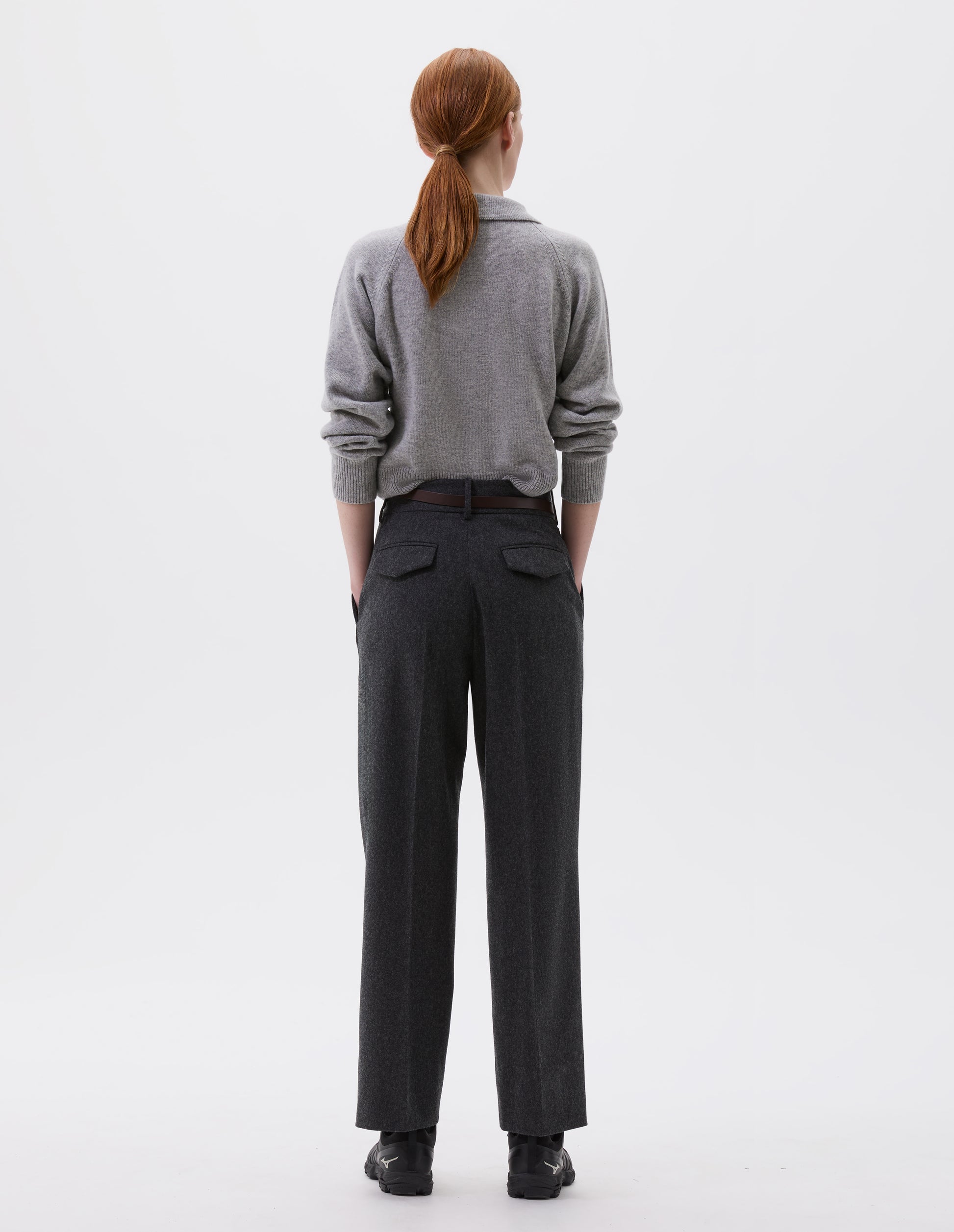 Full body image, back view of model wearing charcoal grey wool trousers with slant side pockets, belt loops and flap back concealed pockets. Styled with grey collared jumper, brown slim leather belt and black trainers.