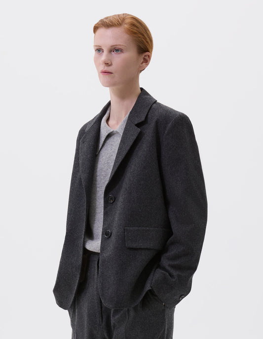 Cropped body image, front view of model wearing dark grey blazer with horn buttons on lapel and cuffs and flap patch pockets. Styled with matching trousers and over a grey collared jumper.