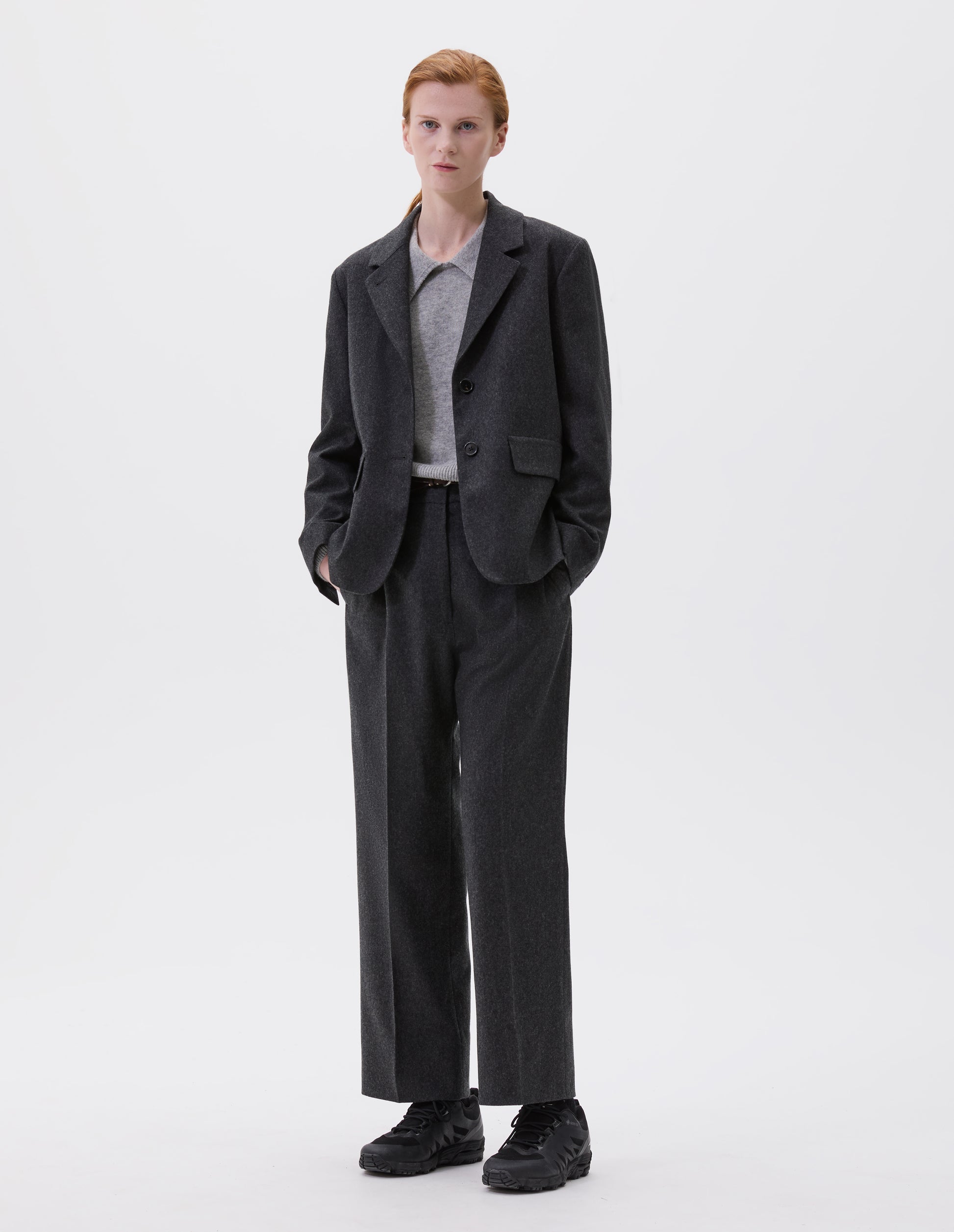 Full body image, front view of model wearing dark grey blazer with horn buttons on lapel and cuffs and flap patch pockets. Styled with matching trousers, over a grey collared jumper and black trainers.