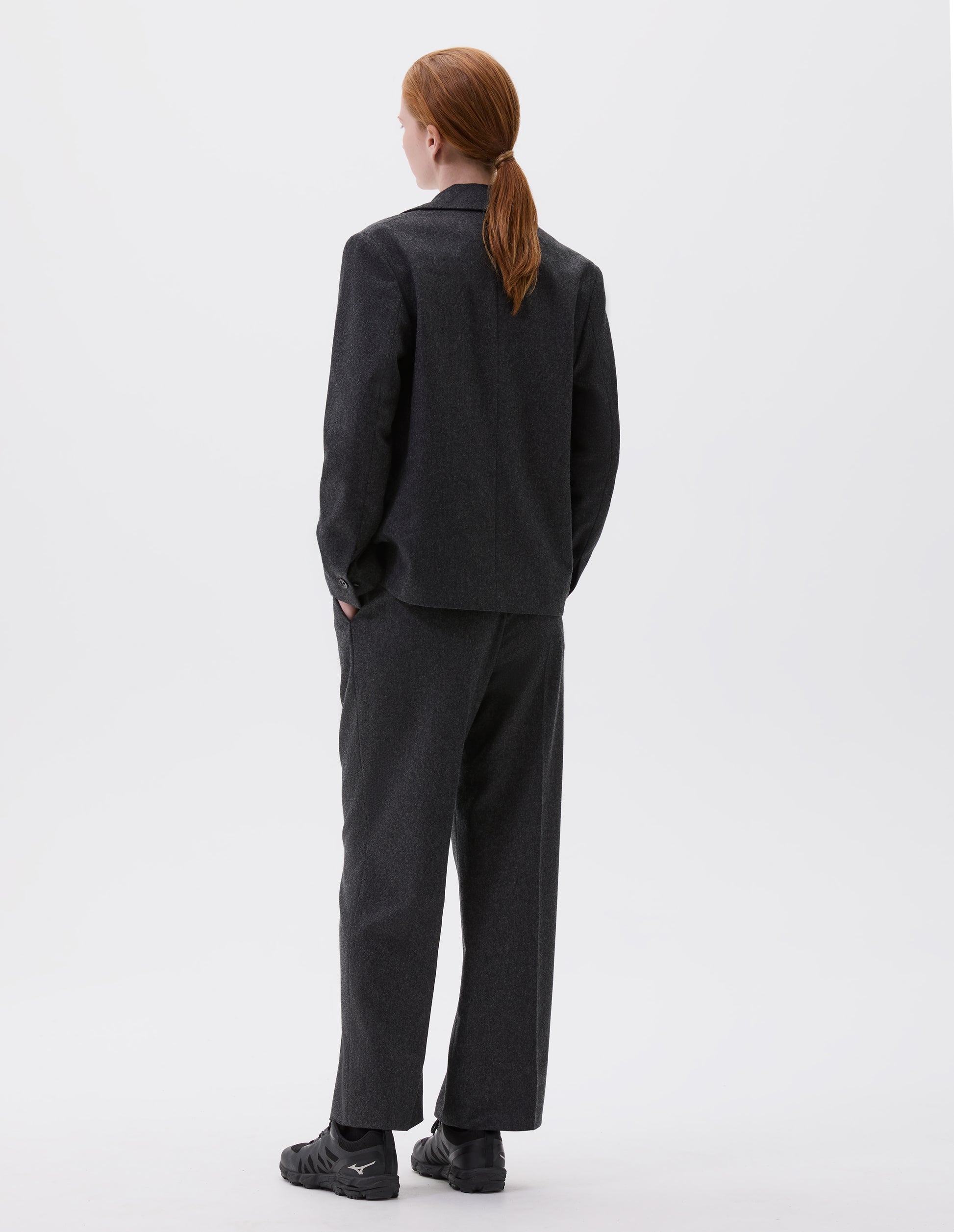 Full body image, back view of model wearing dark grey blazer with horn buttons on cuffs. Styled with matching trousers and black trainers. 
