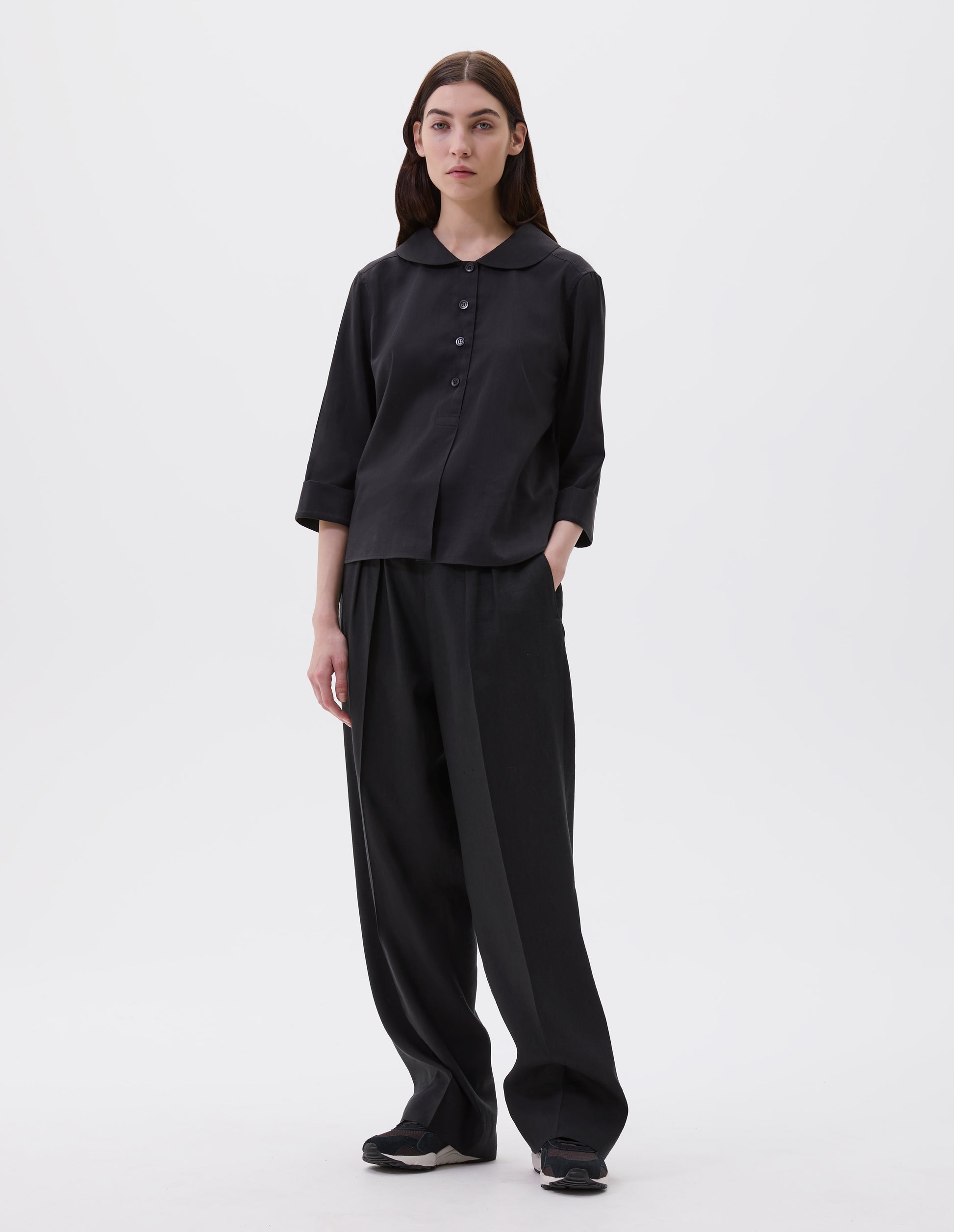 Full body image, front view of model wearing black petal collar shirt with three quarter length sleeves, stitch through half placket, mother of pearl buttons and turned back cuff details. Styled with black pleated trousers and brown and black trainers. 