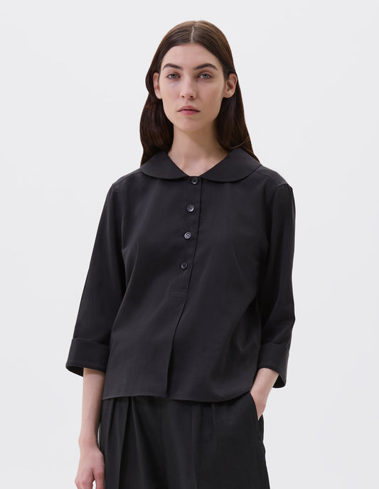 Cropped body image, front view of model wearing black petal collar shirt with three quarter length sleeves, stitch through half placket, mother of pearl buttons and turned back cuff details. Styled with black pleated trousers.