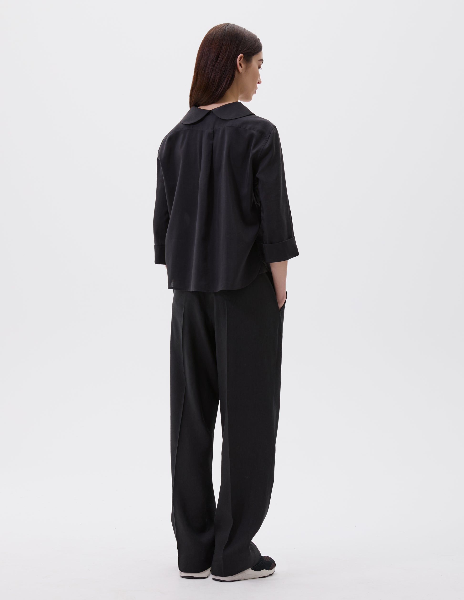 Full body image, back view of model wearing black petal collar shirt with three quarter length sleeves, turned back cuff details and box pleat on back yoke. Styled with black trousers and black and brown trainers.