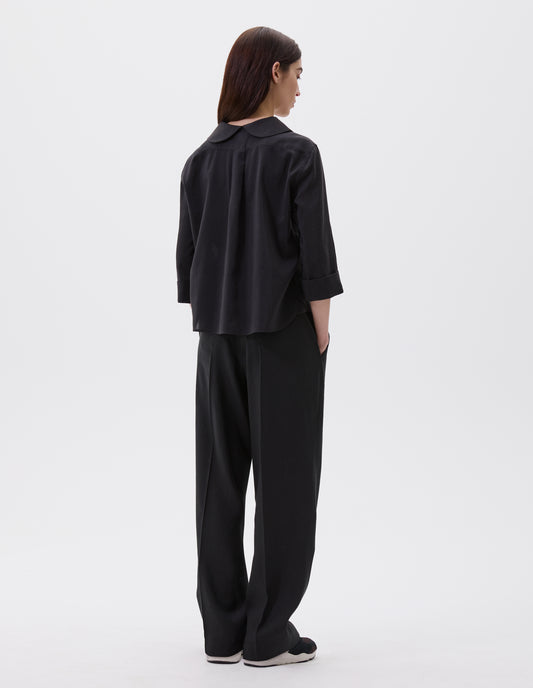 Full body image, back view of model wearing black petal collar shirt with three quarter length sleeves, turned back cuff details and box pleat on back yoke. Styled with black trousers and black and brown trainers.