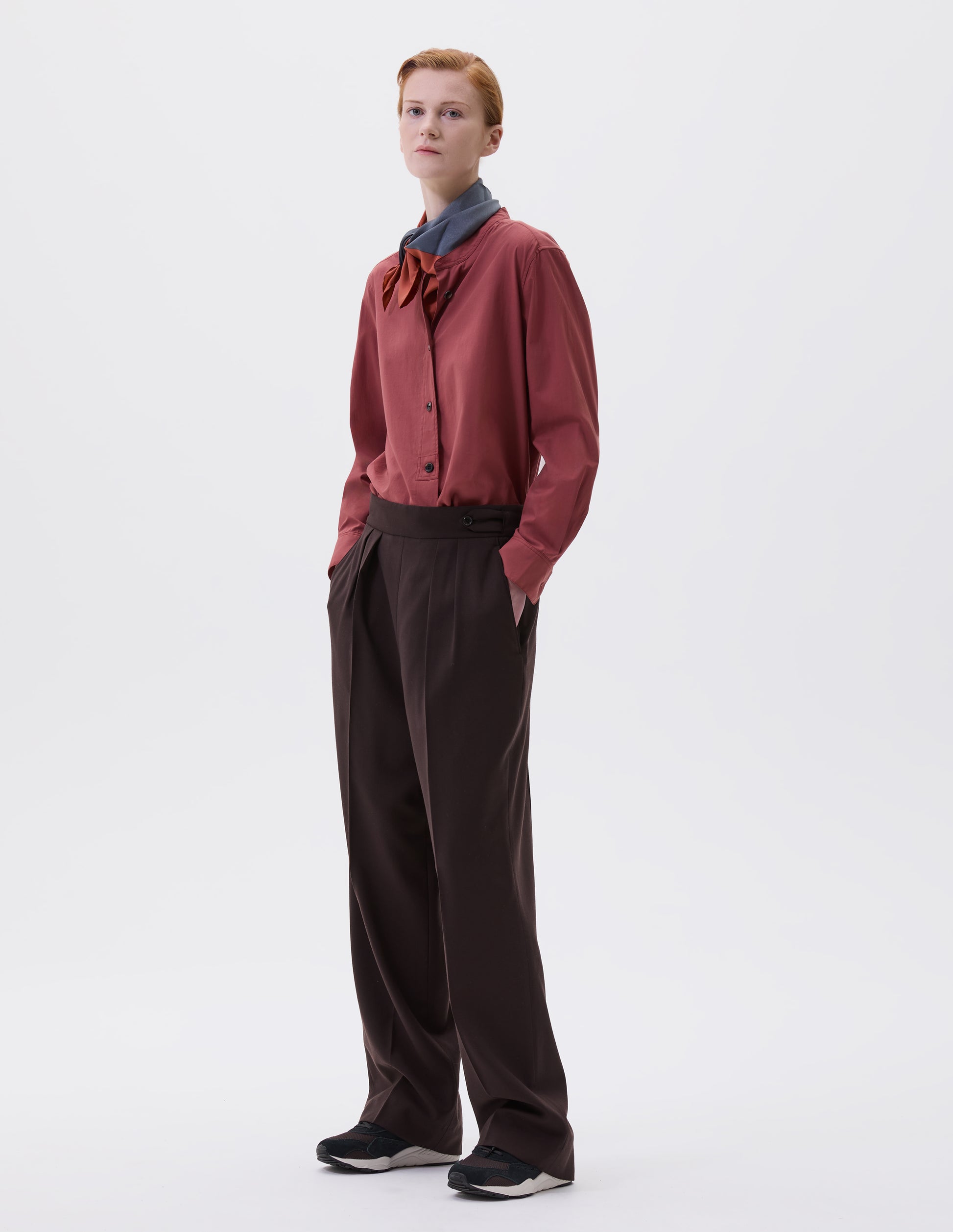 Full body image, front view of model wearing brown double pleated wool trousers, with concealed side zip, pull through tab side fastening and slant pockets. Styled with red shirt, red and blue scarf and black and brown trainers.