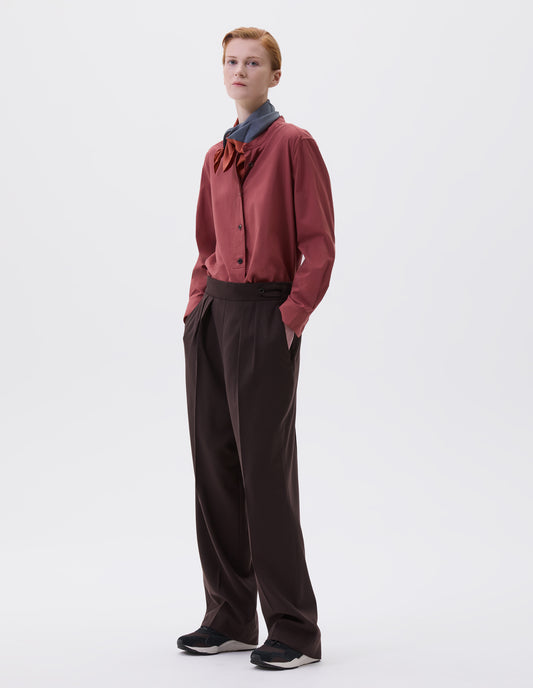 Full body image, front view of model wearing brown double pleated wool trousers, with concealed side zip, pull through tab side fastening and slant pockets. Styled with red shirt, red and blue scarf and black and brown trainers.