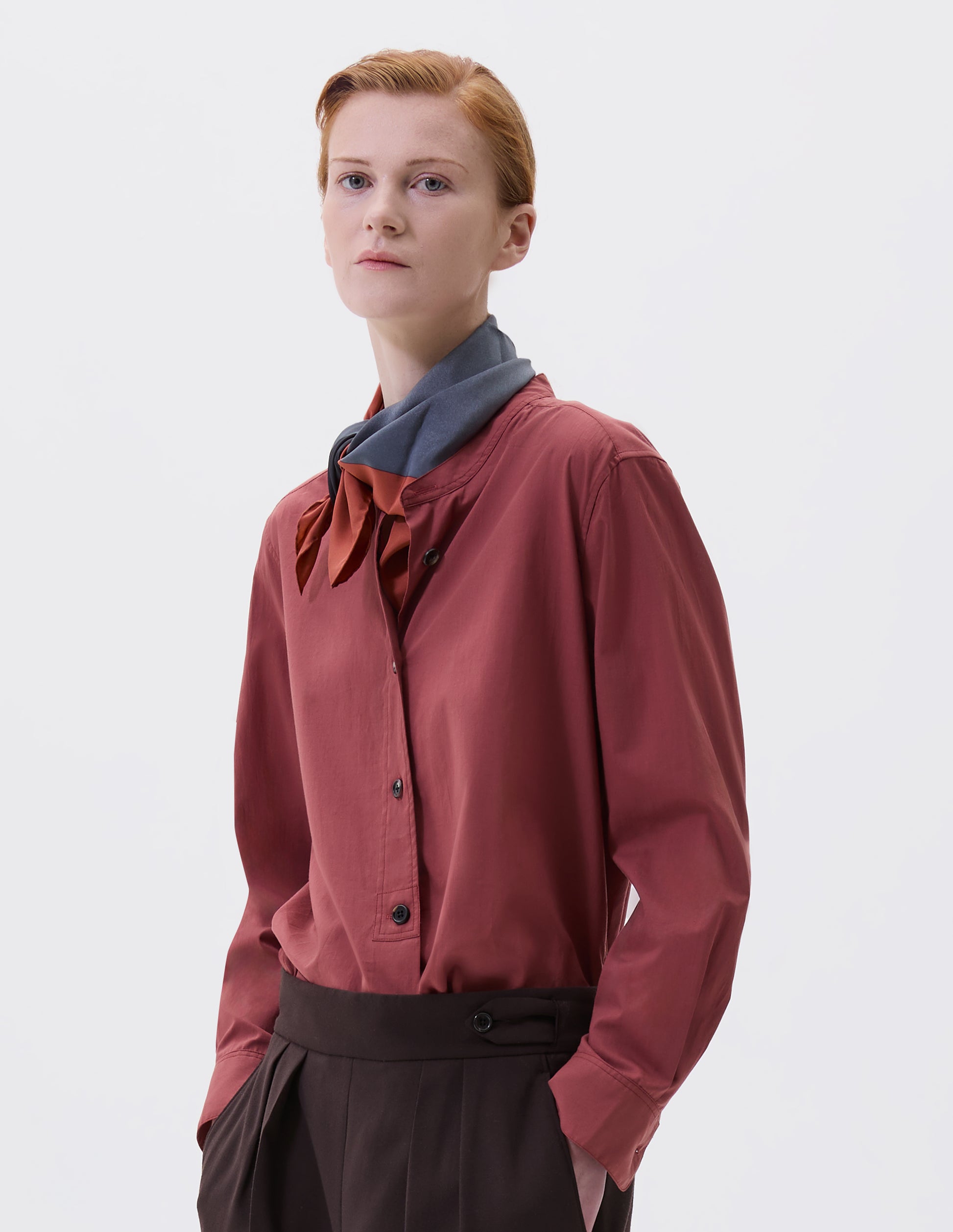 Cropped image, front view of model wearing orange-red tied neck scarf with contrast blue print. Styled with dark pink collarless shirt and brown pleated trousers.