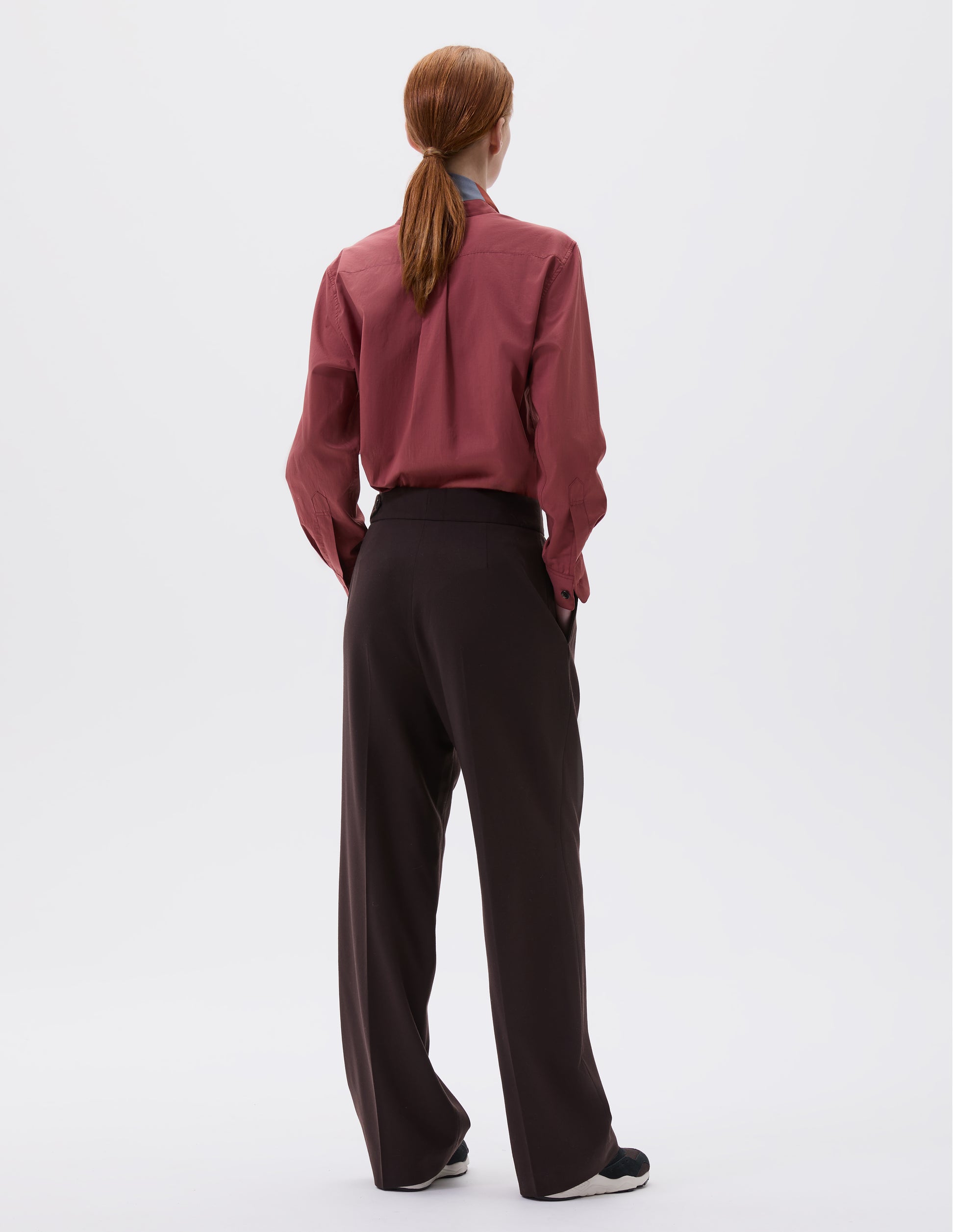 Full body image, back view of model wearing brown double pleated wool trousers, with concealed side zip and pull through tab side fastening. Styled with red shirt, red and blue scarf and black and brown trainers.
