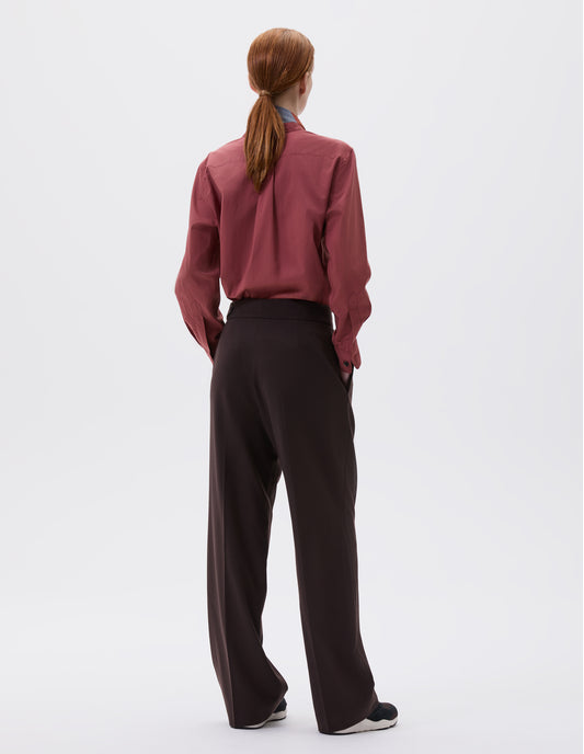 Full body image, back view of model wearing brown double pleated wool trousers, with concealed side zip and pull through tab side fastening. Styled with red shirt, red and blue scarf and black and brown trainers.