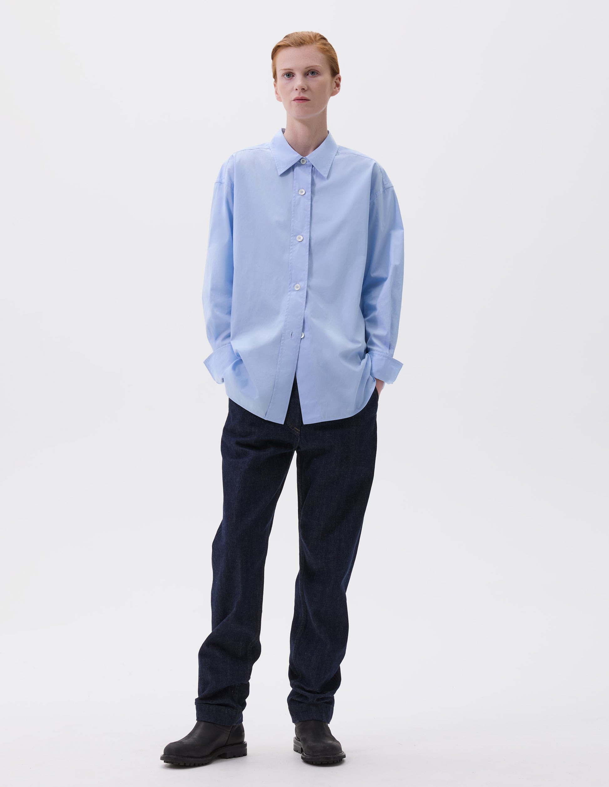 Full body image, front view of model wearing relaxed light blue shirt with wide raised placket and mother of pearl buttons, and turned up cuffs. Styled with dark blue jeans and black leather boots. 