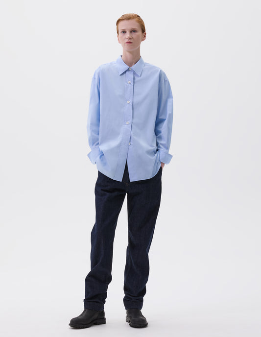 Full body image, front view of model wearing relaxed light blue shirt with wide raised placket and mother of pearl buttons, and turned up cuffs. Styled with dark blue jeans and black leather boots. 