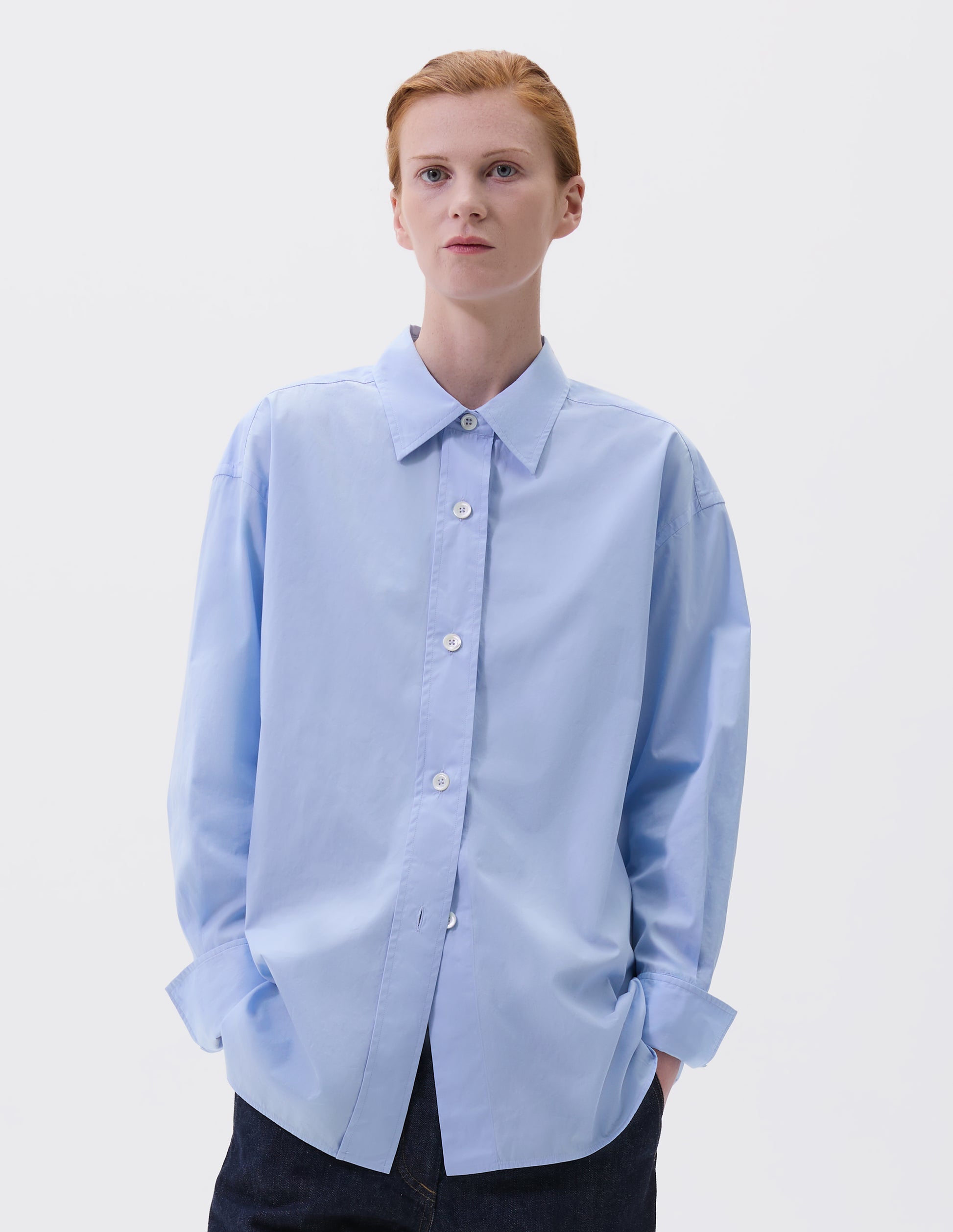 Cropped image, front view of model wearing relaxed light blue shirt with wide raised placket and mother of pearl buttons, and turned up cuffs. Styled with dark blue jeans.
