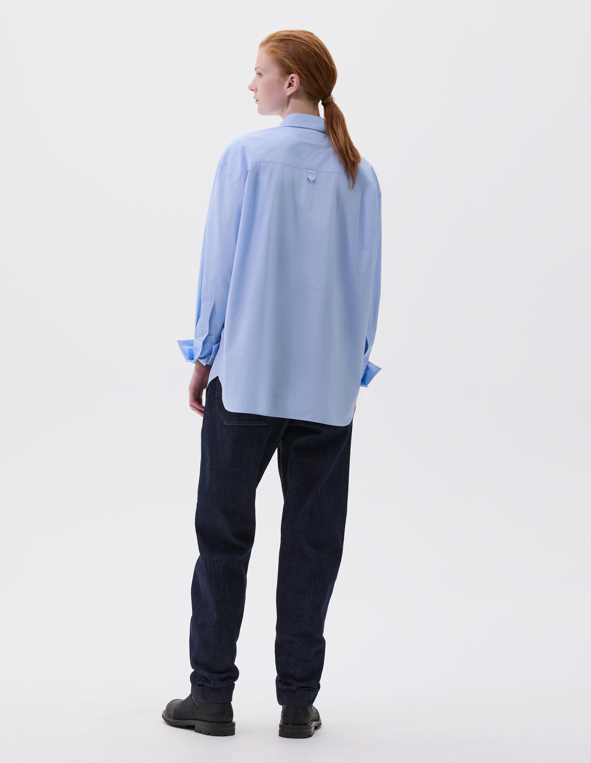 Full body image, back view of model wearing relaxed light blue shirt with mother of pearl buttons on turned up cuffs. Styled with dark blue jeans and black leather boots.